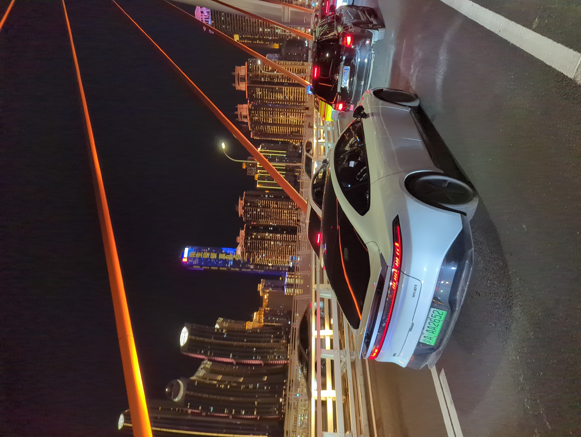 The image shows a nighttime scene on a bridge in Yuzhong District, China. The bridge is illuminated by orange lights that run along its cables and structure. The background features a dense cityscape with numerous tall buildings, many of which are lit up. Several cars are visible on the bridge, suggesting traffic. In the foreground, a white electric vehicle with a distinctive taillight design is prominent. Another vehicle, a dark SUV, is behind it. The road surface is asphalt, marked with white lines. The sky is dark and clear, with no visible celestial bodies.