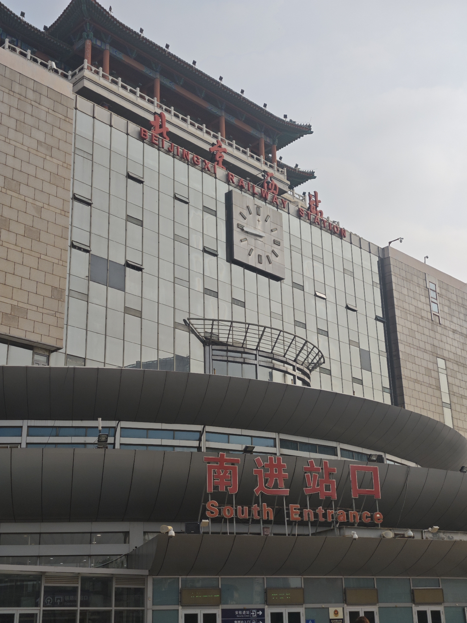 The image is a medium shot of the Beijingxi (West) Railway Station in the Fengtai District of Beijing, China. The station's modern facade features a grid of glass windows, a large clock, and red signage in both Chinese characters and English that reads "BEIJINGXI RAILWAY STATION." Below this, a curved gray structure displays the words "South Entrance" in English and Chinese. The base of the building houses the station's entrance with visible doors, signage, and lighting fixtures. The sky is overcast, and the image is taken from a low angle, emphasizing the building's height. The style of the station incorporates traditional Chinese architectural elements, evident in the tiered roof structure above the modern building. A sign in Chinese and English reads, "Security Check" and shows the symbol for people with disabilities.