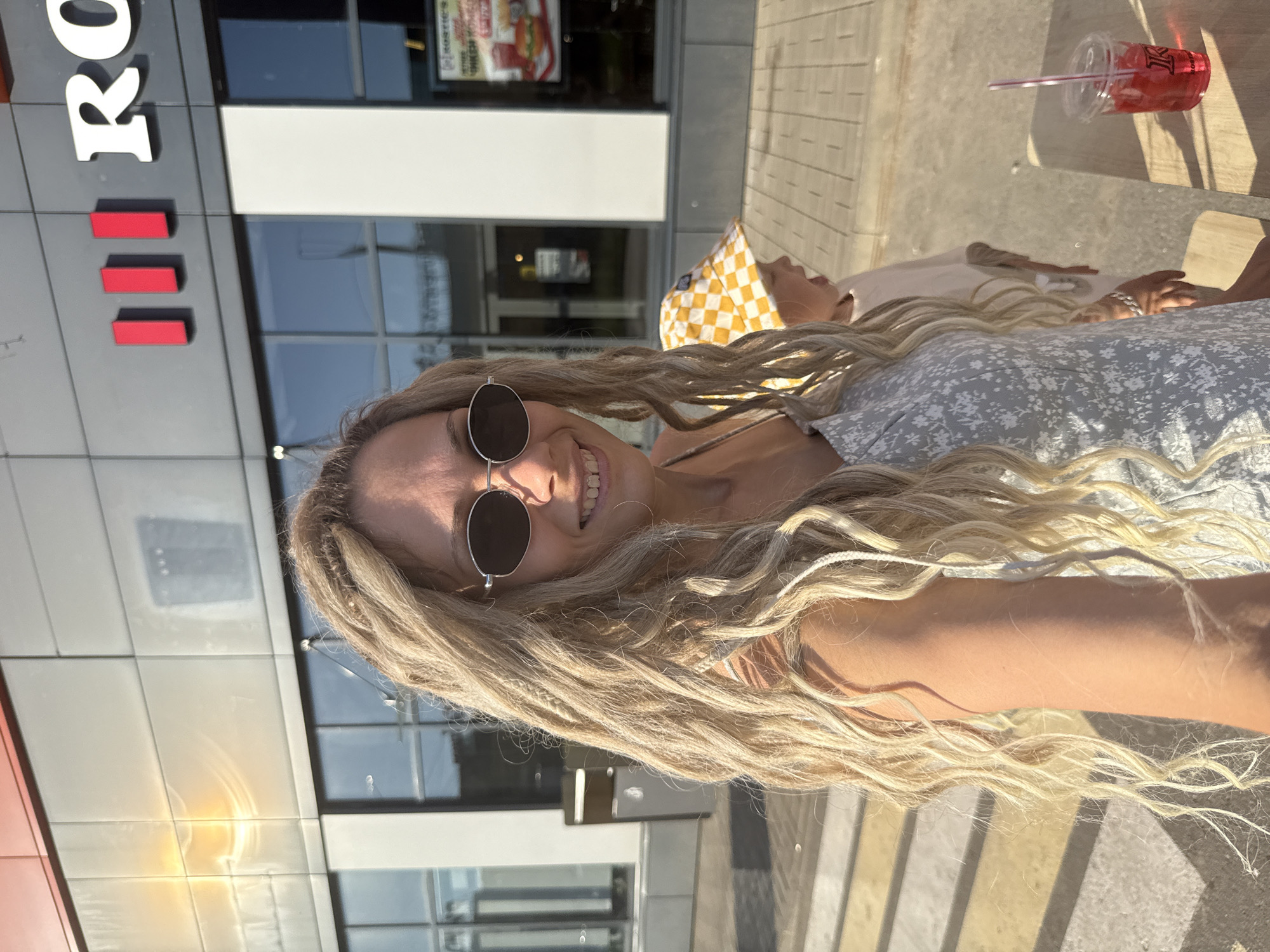 The image depicts a woman with blonde dreadlocks, wearing sunglasses and a light-colored floral dress, posing for a selfie outdoors. Behind her, a young child wearing a checkered hat is visible. The background features a building with large windows and signage that appears to be in Russian, suggesting an urban setting. The lighting indicates it is daytime. There is a red drink in a clear cup with a straw on a wooden surface in the foreground. The presence of the building and signage could indicate a commercial establishment. The overall scene suggests a casual outing in an urban environment.