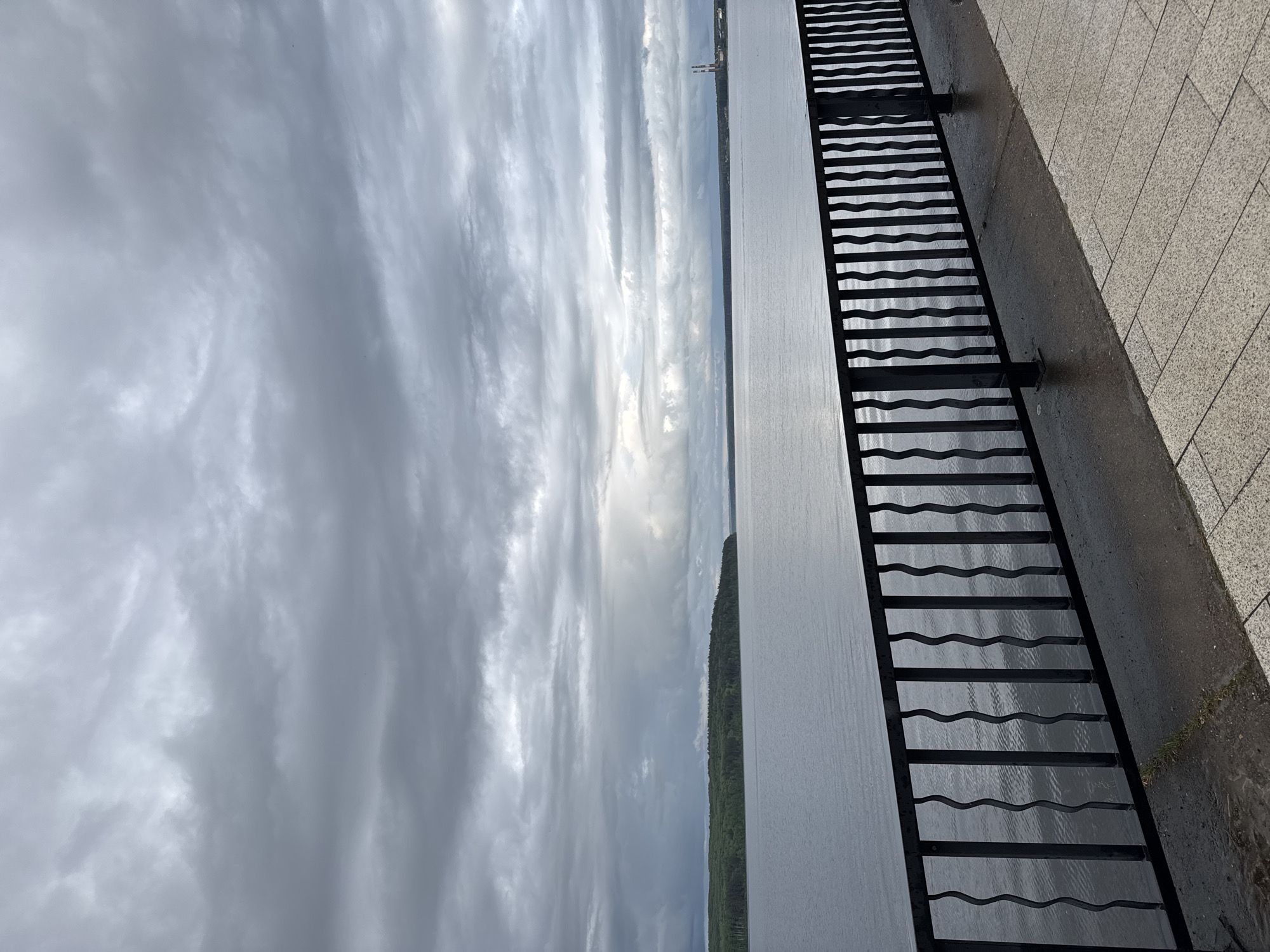 The image, rotated 90 degrees clockwise, depicts a wide landscape scene from what appears to be a waterfront promenade. When viewed upright, a large expanse of water occupies the middle ground, stretching horizontally across the frame. Its surface is subtly textured with ripples, reflecting the cloudy sky above.
The sky, filling the upper portion of the image, is heavily overcast with dramatic bands of grey and white clouds. A prominent bright area of light breaks through the clouds in the distant center, suggesting the presence of the sun or a bright patch of sky beyond the immediate cloud cover.
On the far bank of the water, a continuous line of dark green forest marks the horizon. Above this tree line, two light-colored industrial structures, resembling tall chimneys or towers, are visible against the distant sky.
In the foreground, running along the right side of the image, is a dark metal railing with a distinctive design of horizontal, wavy bars. This railing is mounted on a darker, possibly concrete, wall or base, which shows support brackets. Adjacent to this base, on the far right, is a light-colored paved embankment or walkway made of square tiles. Both the tiled surface and the concrete base appear damp, suggesting recent rain.
There are no people or specific activities taking place. The overall atmosphere is serene and open, under an overcast sky with distant light. No text is visible in the image. The scene suggests a large river or reservoir, consistent with the geographic context of Chaikovsky, Russia.