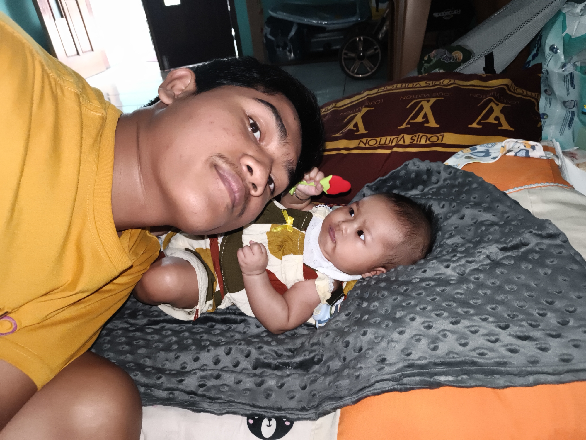 A young man, possibly in his late teens, lies on his back, wearing a bright yellow t-shirt. He is looking directly at the camera, with a faint smile and a mustache. Next to him, also on his back, is a baby wearing a patterned outfit in shades of brown, yellow, and white, with a white bib. The baby is looking upwards, with a toy in its right hand. They are lying on a gray, textured blanket. Behind them, a patterned pillow with "LOUIS VUITTON" visible is present. The environment appears to be indoors, possibly a bedroom, with hints of furniture and a door in the background. The scene is likely located in Barengkrajan, Indonesia, as per the prompt, though no specific landmarks are visible to confirm this.