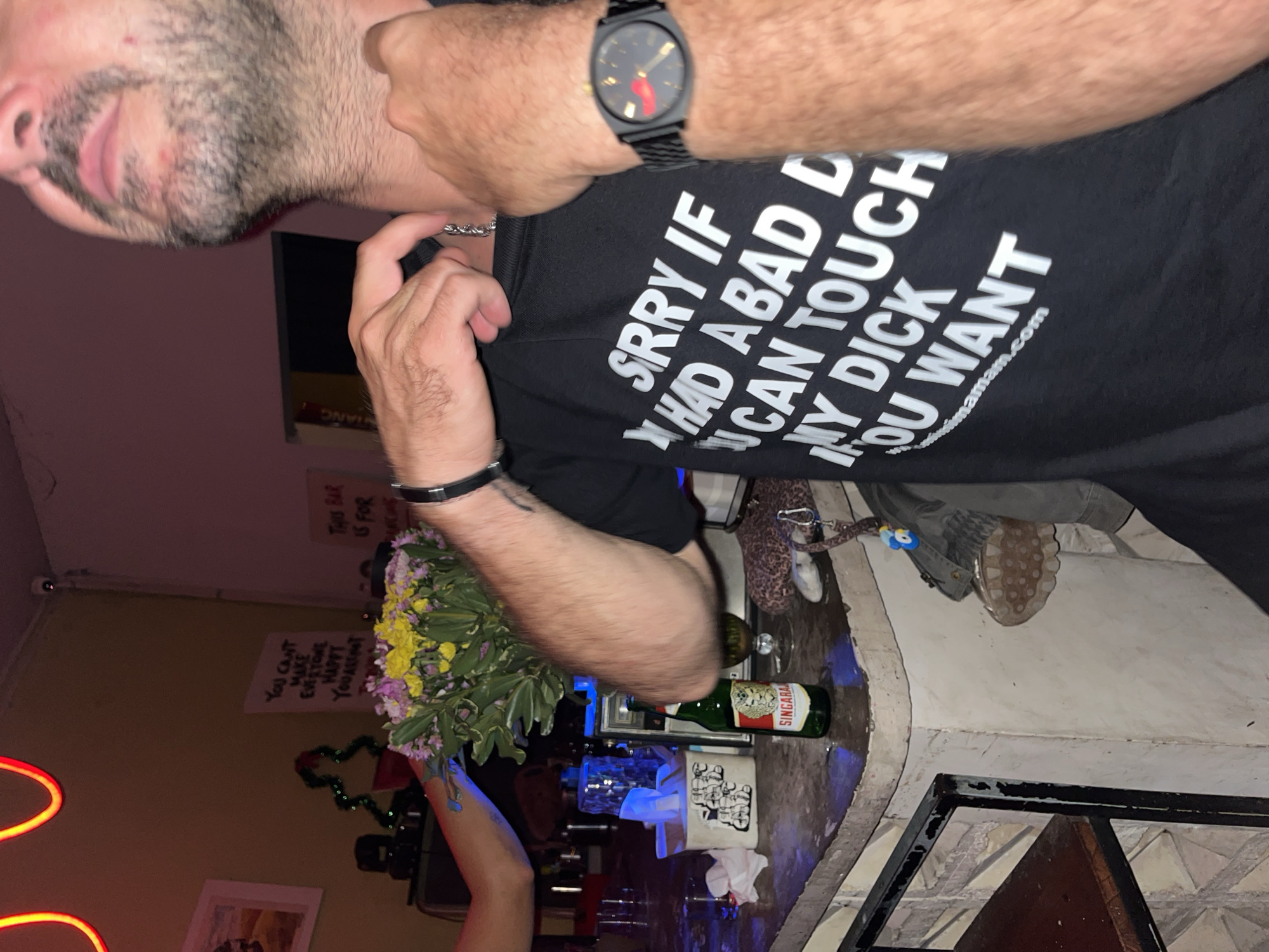 A man is wearing a black t-shirt with a sexually suggestive message. The text on the shirt reads, "SRRY IF I HAD A BAD D I CAN TOUCH MY DICK IF YOU WANT." He is also wearing a black watch and a silver chain. He is standing next to a bar counter with a bottle of Singha beer and a bouquet of flowers. The background includes bar decor, signs, and neon lights. The location appears to be in North Kuta, Indonesia.