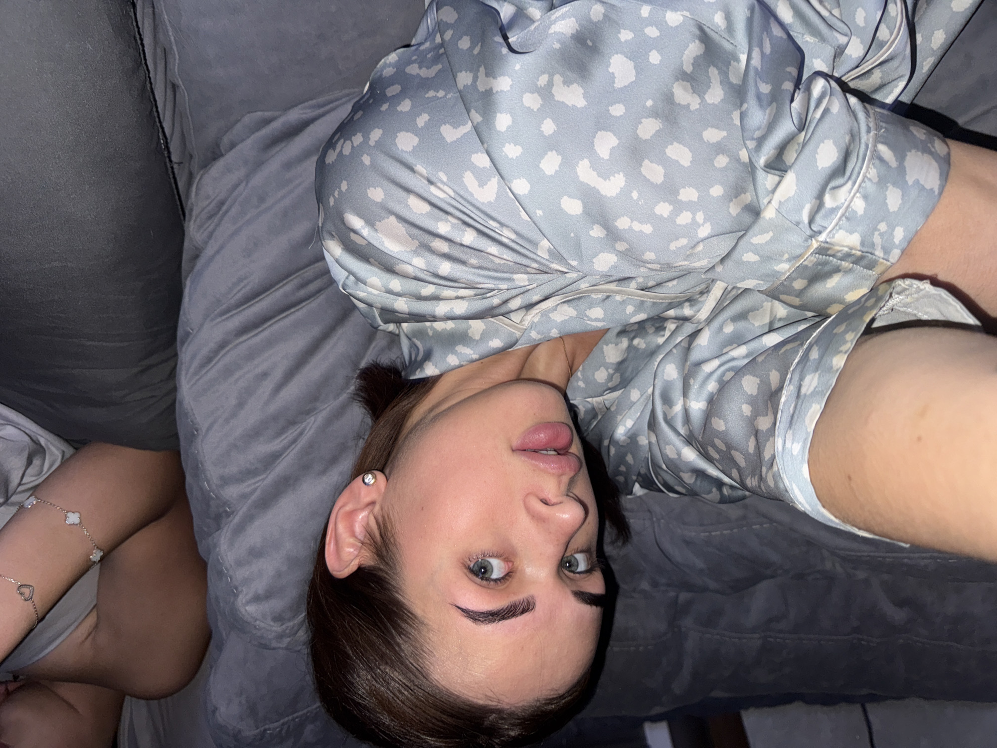 A woman lies on a grey sofa, possibly in a home. She is wearing a light blue patterned top. Her left arm, adorned with a bracelet, is visible at the bottom left of the image. Her head is tilted, and she looks into the camera. The location of Лобаново, Russia, is not discernible from the image.
