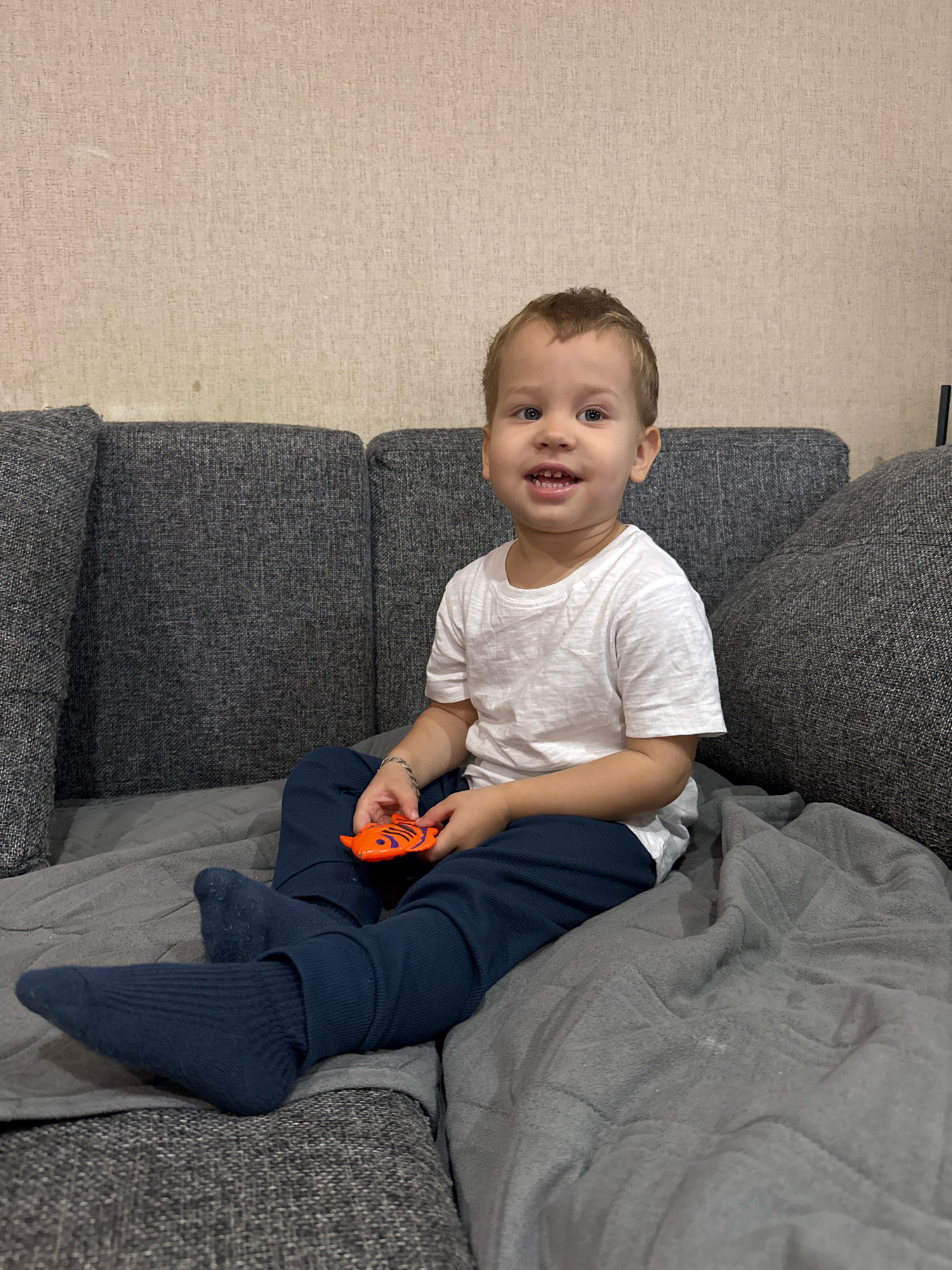 A young Caucasian boy, approximately 2-3 years old, is seated on a gray sofa with a textured, light-colored wall behind him. The boy has light brown hair and blue eyes, and he is wearing a white t-shirt, navy blue pants, and navy blue socks. He is holding an orange toy shaped like a fish in his right hand. The sofa is covered with a gray blanket. The lighting appears to be artificial, suggesting the image was taken indoors. The overall environment suggests a casual, domestic setting. The location is identified as Rusanovo, Russia.