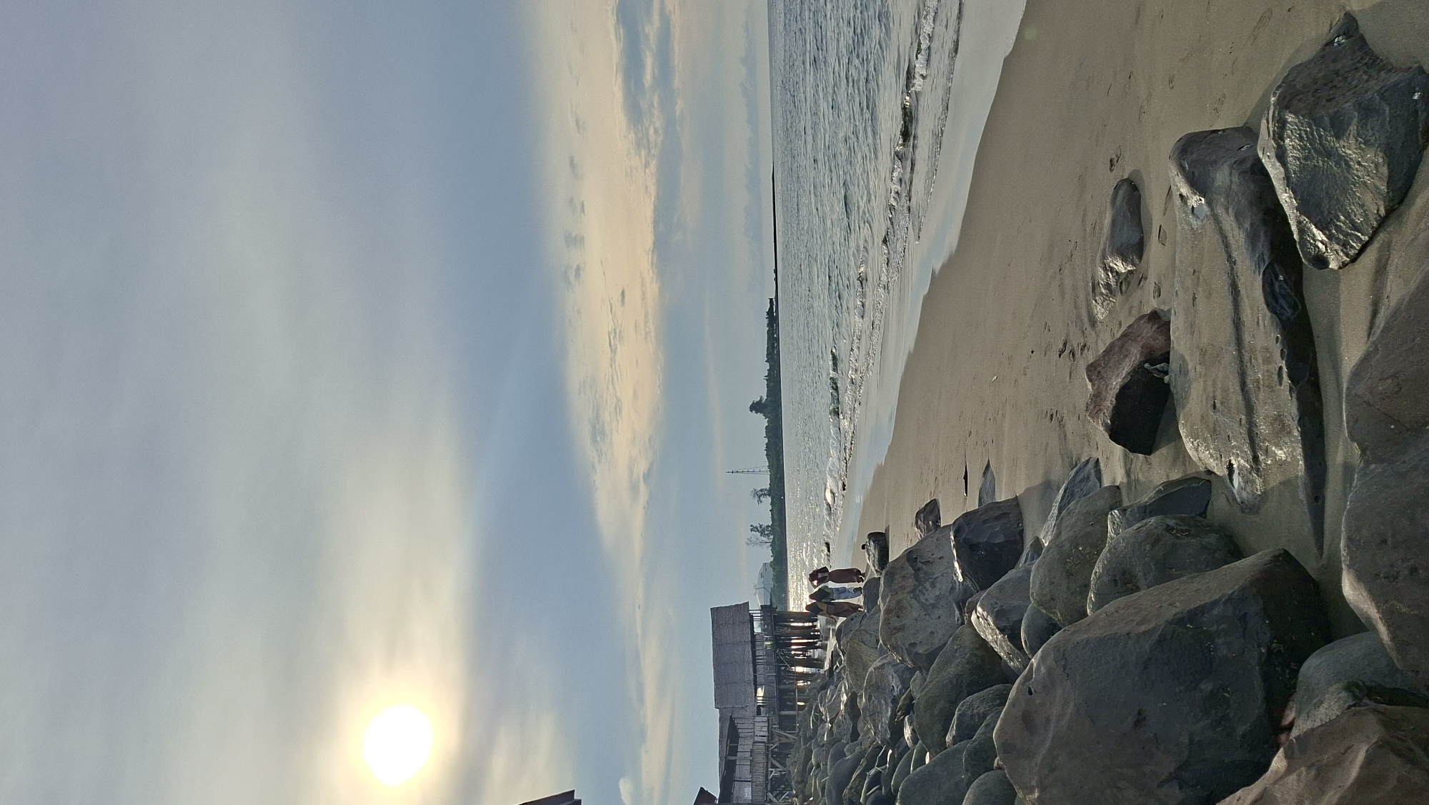 This is a landscape photograph of a beach in Lhokseumawe, Indonesia, taken at sunset. The sun, a bright orb, is low in the sky and casts a warm glow, illuminating the scene with soft light. The sky is a pale blue, streaked with wispy clouds that are tinged with yellow and orange from the setting sun.
The foreground is dominated by a line of large, dark rocks that stretch along the edge of the sandy beach. The sand itself is a light tan color, showing the gentle ebb and flow of the tide. The ocean is calm, with small waves lapping at the shore.
In the middle ground, there are some stilt houses, appearing to be made of wood and straw, built close to the water's edge. A few people are visible near these structures, though their features are not distinct. Their presence suggests a relaxed, perhaps leisurely, atmosphere.
Further in the distance, along the coastline, a thin line of trees marks the land. A tall structure, possibly a tower or antenna, is also visible on the horizon. The overall mood of the image is serene and peaceful, capturing the tranquil beauty of a tropical sunset. The scene is illuminated by the low sun, creating a warm and inviting ambiance. There is no visible text in the image.