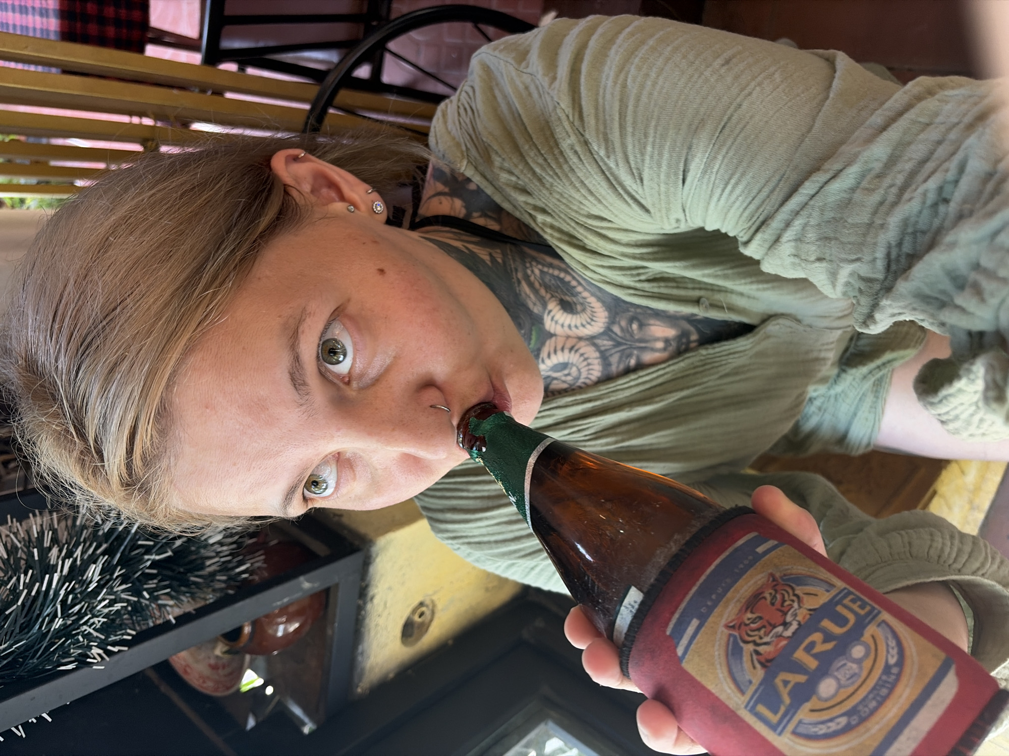 A close-up, angled portrait features a person drinking from a beer bottle. The subject has light blonde or light brown hair and green or hazel eyes that gaze directly at the camera. They sport multiple ear piercings, including hoops, studs, and a decorative cluster, as well as a silver hoop nose piercing. Intricate blackwork tattoos are visible on their neck, shoulder, and upper chest. They are dressed in a loose, light olive green long-sleeved top with a crinkled texture, with a black strap visible underneath.
The person is shown with their lips around the neck of a brown glass beer bottle, which is partially covered by a red fabric cozy. The cozy is adorned with an illustration of a tiger's head and the prominent brand name "LARUE." The background suggests an indoor or covered outdoor setting, possibly a cafe or bar, with wooden slats or beams overhead and part of a black metal chair frame visible. To the left, a dark, spiky decorative item rests on a shelf alongside a reddish-brown pot. The scene is illuminated by natural light, indicating it is daytime. The subject's expression is direct and engaged. This scene is set in Hội An Ward, Vietnam.