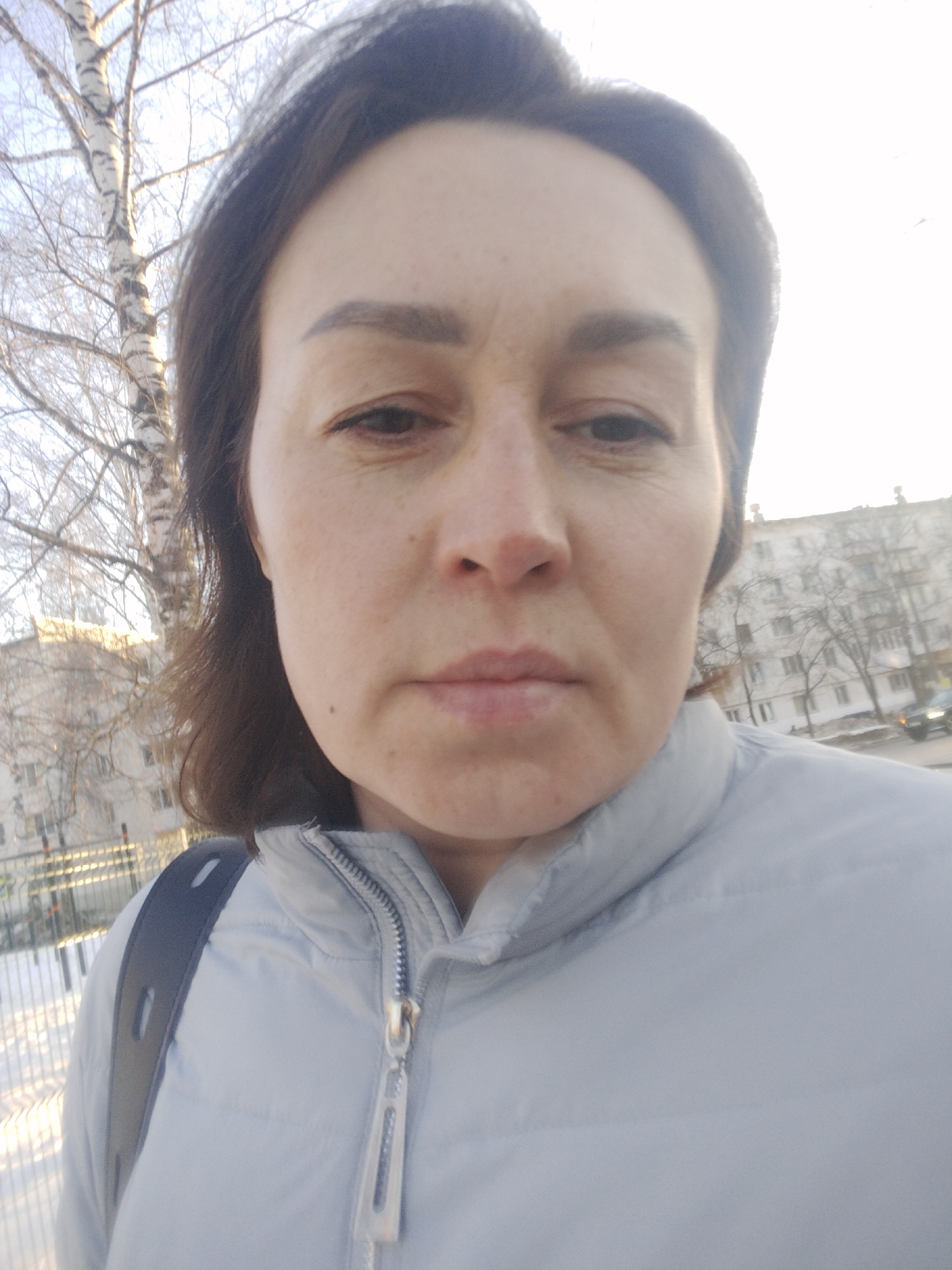A close-up photograph shows a middle-aged woman with brown hair and a light skin tone wearing a light-blue quilted jacket. She is looking directly at the camera with a neutral expression. A dark-colored strap, likely belonging to a backpack, is visible over her left shoulder.
The background reveals an urban environment, identified as the city of Chaikovsky, Russia. A bare birch tree stands on the left side of the frame. In the distance, several multi-story buildings are partially visible, with bare trees lining the street. There is snow on the ground. The lighting suggests it is daytime.