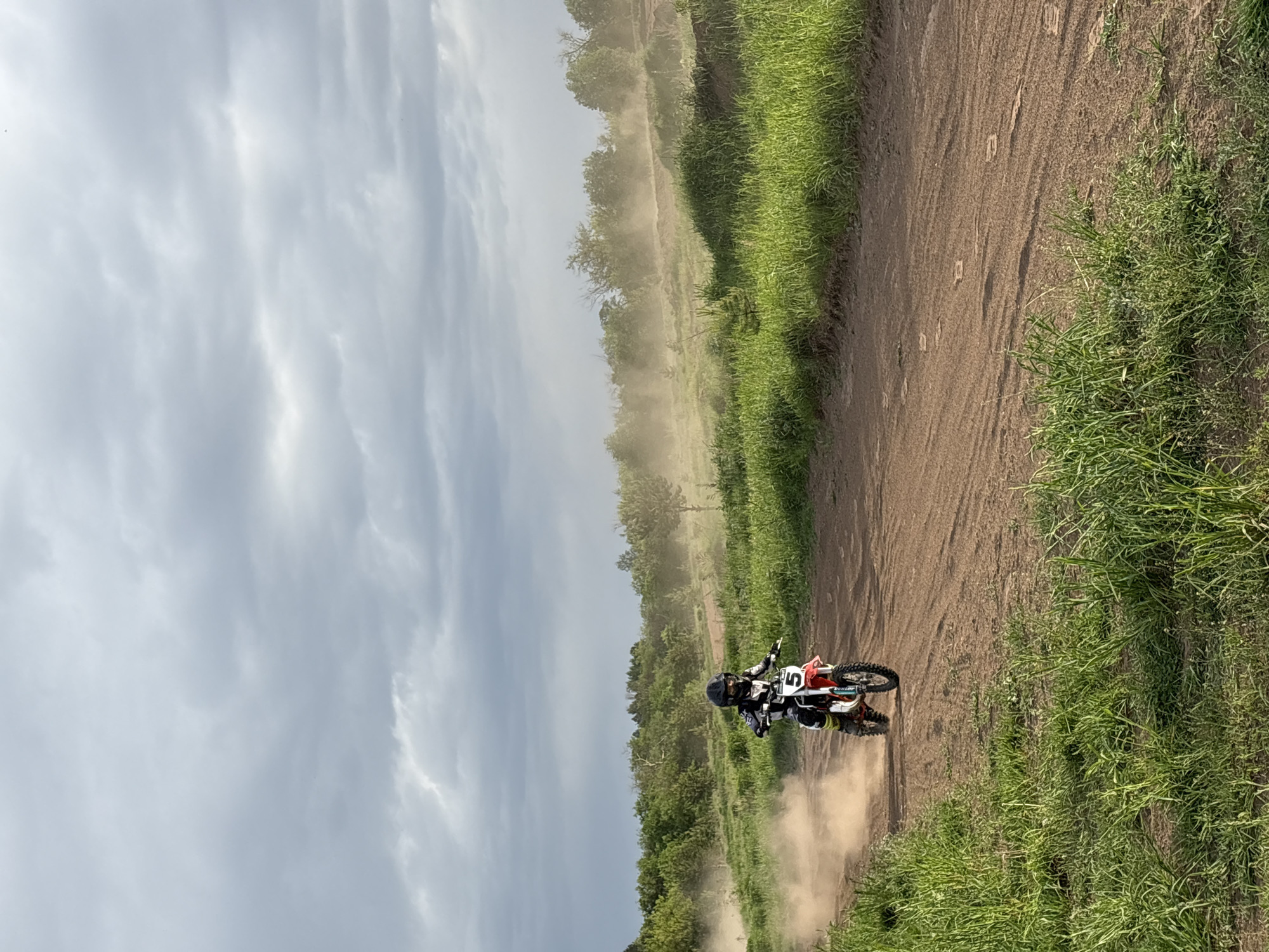 A motorcyclist is riding a dirt bike on a dirt road. The road is bordered by green vegetation on the right and trees in the distance on the left. The sky is overcast. The motorcycle is kicking up dirt as it moves along the road. The image is rotated 90 degrees counterclockwise. It is not possible to determine the location in Votkinsk, Russia from the image.
