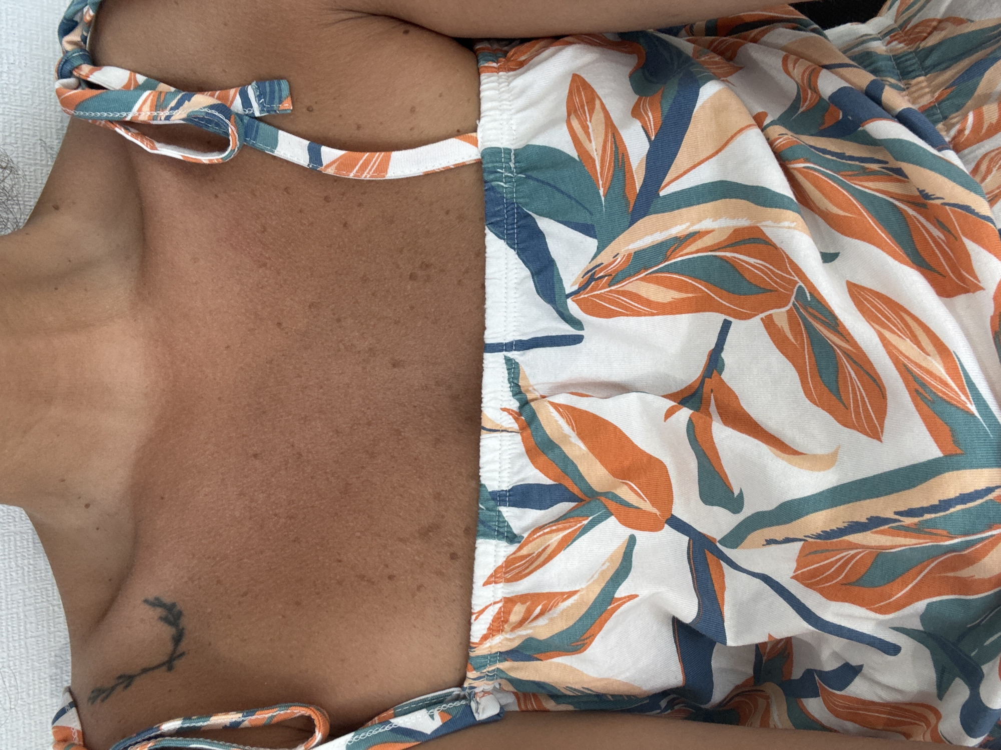 The image shows a close-up of a person's upper chest and shoulders, wearing a white top with a tropical leaf pattern in orange, teal, and blue. A small tattoo of a sprig is visible on the left shoulder. The skin appears tanned and has freckles. The background is a plain, textured surface, likely a wall or fabric. The image is cropped tightly on the subject and does not provide any context about the location within the city of Khashuri, Georgia.