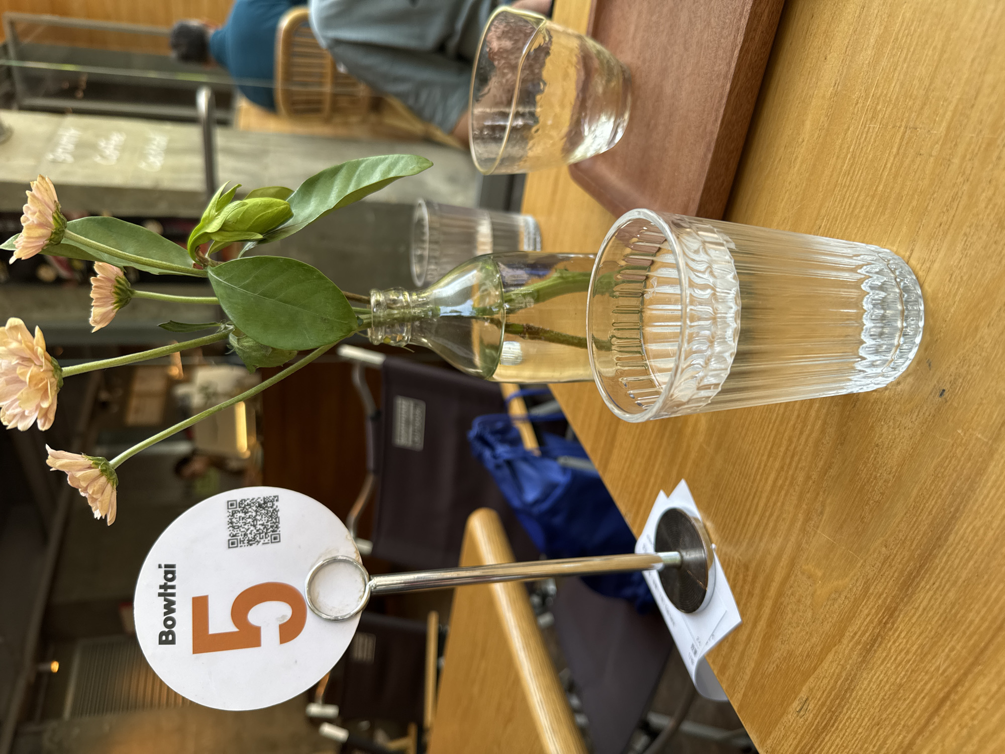 The image depicts a table setting in a restaurant, potentially located in the Yuzhong District of China, based on the "Bowltali" brand name on the table number. The table is made of wood and has a table number of "5" displayed on a white, circular sign with a brown number and QR code. A small glass vase with flowers is in the center of the table, along with two drinking glasses. The background includes other tables, chairs, and people, suggesting a busy restaurant environment. A blue bag is also visible, likely belonging to a customer.
