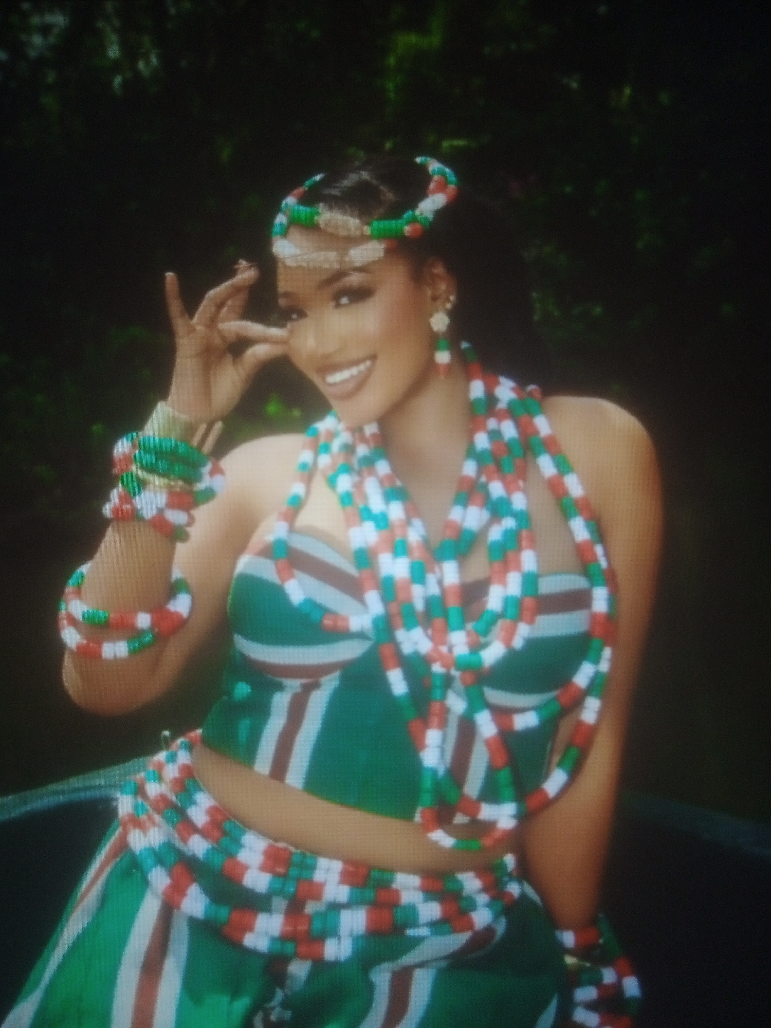 A woman with dark hair and fair skin is posing in front of a dark, leafy background, suggesting an outdoor setting. She is wearing a green and white striped bustier top and a matching skirt. Adorning her are numerous strands of red, white, and green beads, worn as necklaces, bracelets, and a headband. Her right hand is raised near her face, with her fingers delicately extended. She is smiling broadly, showing her teeth.
