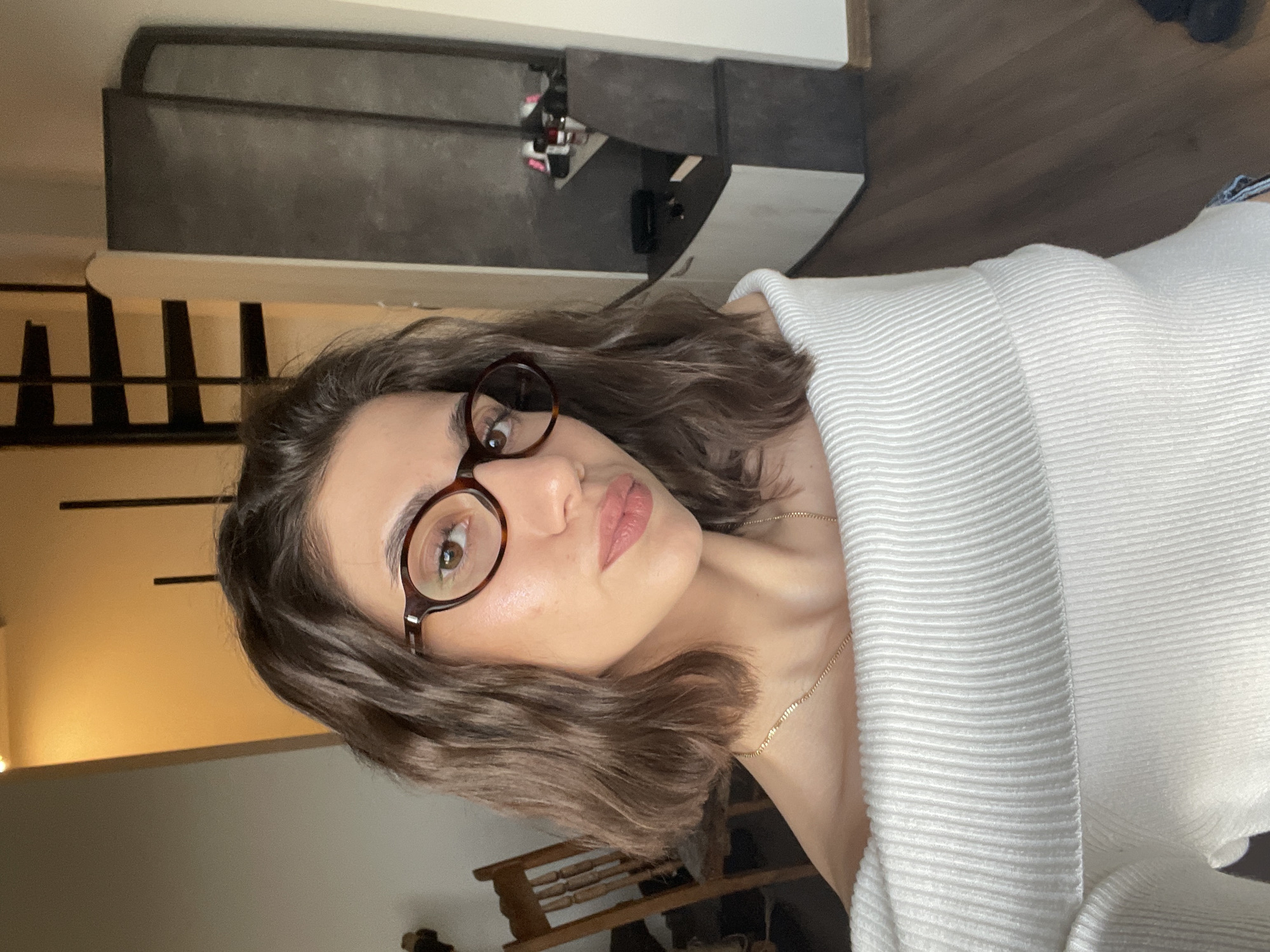 A woman is photographed indoors, wearing brown-rimmed glasses, a white sweater, and a gold chain. She has shoulder-length brown hair. The background includes a staircase with black metal railings, a dark-colored cabinet with various items on top, and wooden flooring. A glimpse of a wooden chair is also visible. The location cannot be confirmed as Sofia, Bulgaria, based solely on the visual information.