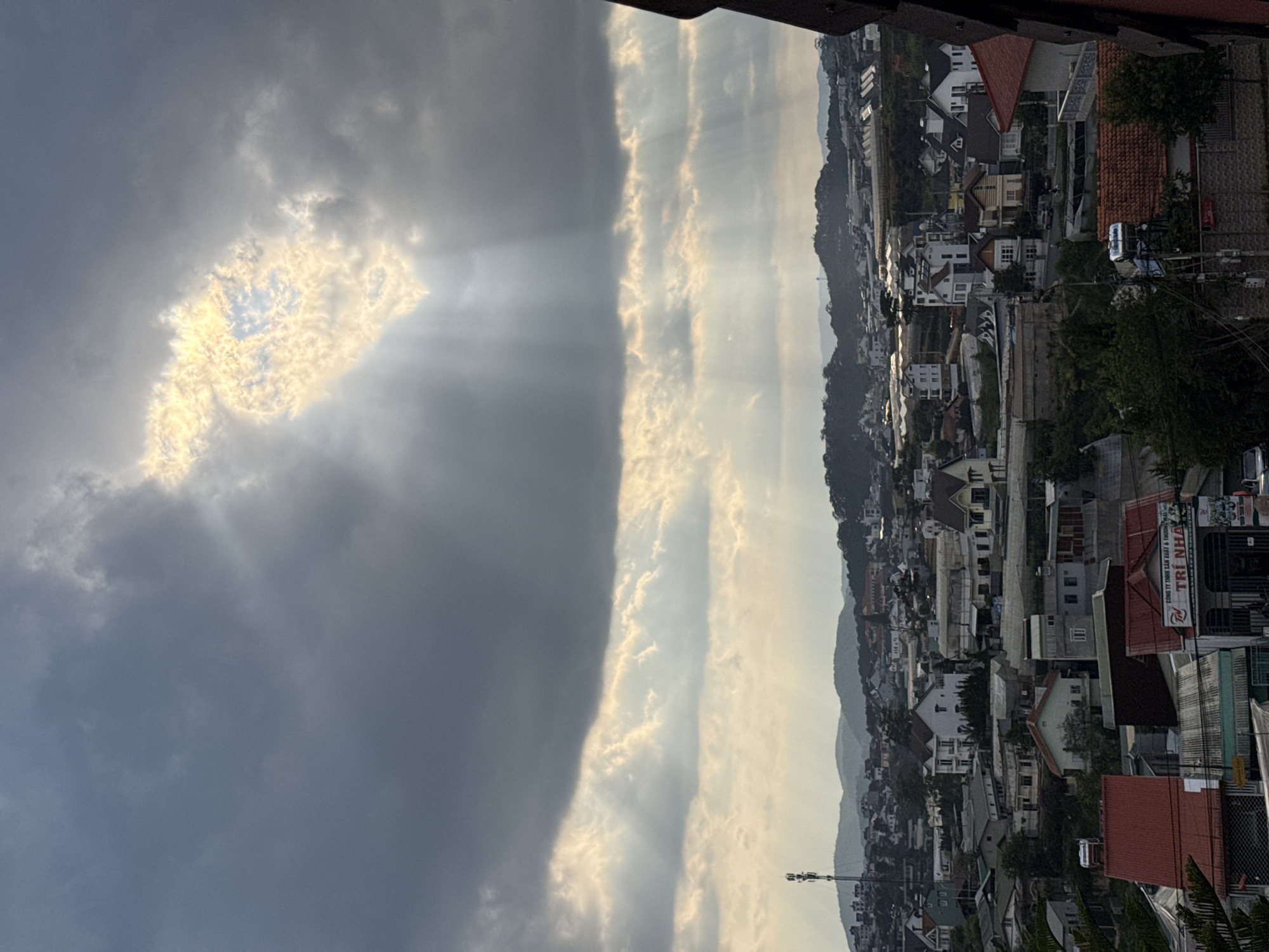 The photograph, taken in Lâm Viên - Đà Lạt, Vietnam, presents a vertical landscape view dominated by atmospheric phenomena. On the left, dark, heavy clouds obscure the sky, save for a radiant, irregularly shaped opening through which the sun bursts, scattering light beams. To the right, the sky transitions to lighter, brighter tones, marked by streaks of sunlight. Below, a dense urban expanse stretches into the distance, characterized by clustered buildings of various sizes and colors, predominantly featuring red and white roofs. The landscape includes rolling hills in the background. A communications tower is visible among the buildings. The composition suggests a moment where a storm is breaking, allowing sunlight to pierce through the cloud cover over a densely populated area.