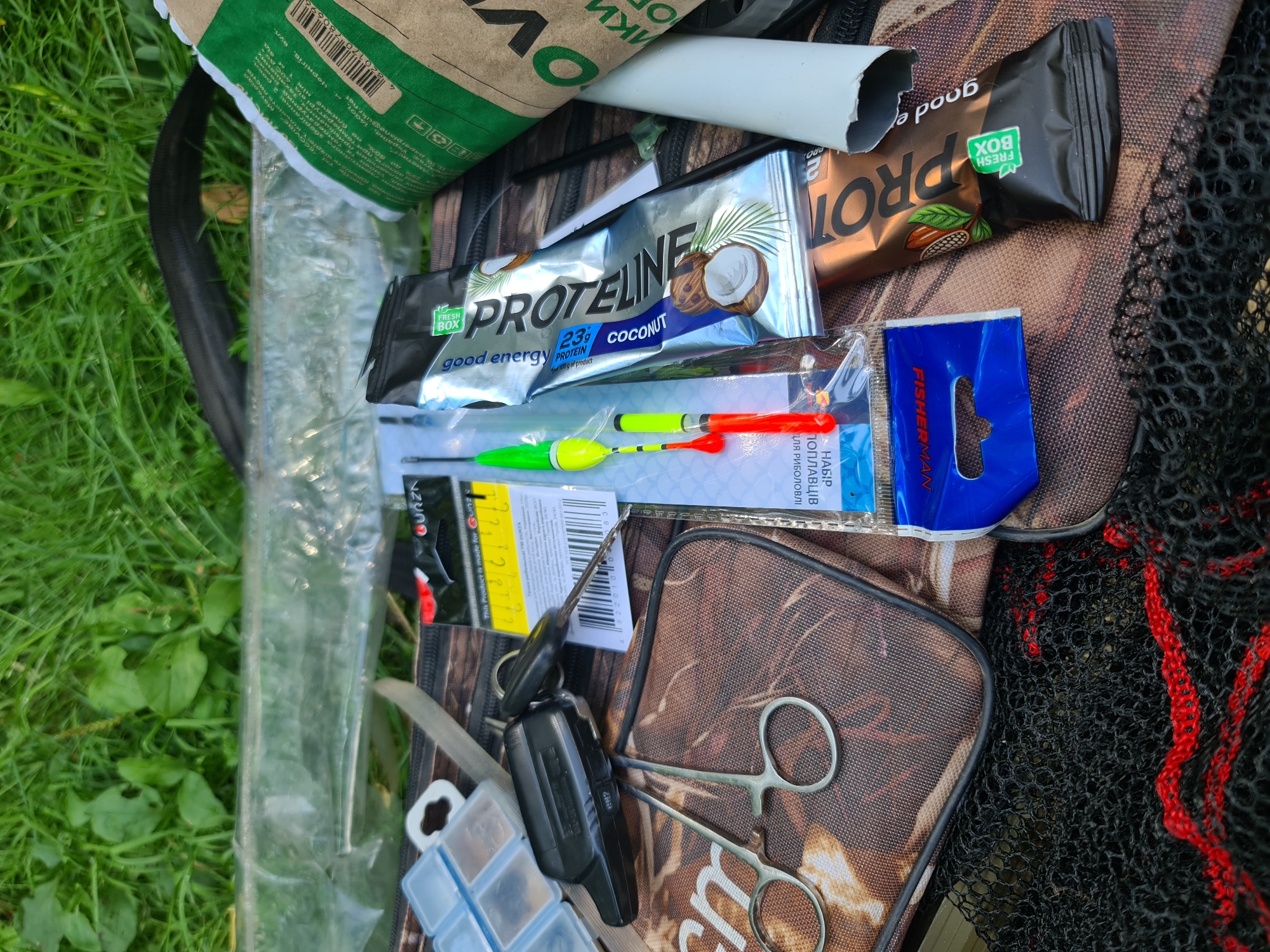 In the city of Potoky, Ukraine, an outdoor scene is depicted, likely a fishing trip, given the presence of fishing tackle. The image shows a collection of items laid out on a camouflage-patterned bag and a black fishing net. Prominently featured are fishing floats, fishing hooks in a clear plastic container, a pair of small scissors, and a set of keys with a car remote. Alongside the fishing gear are two protein bars, one flavored with coconut and another with chocolate. A green and white paper bag with text in Ukrainian is also visible, suggesting a purchase or product packaging. The background consists of lush green grass and a hint of a clear plastic sheet. The lighting suggests it is daytime, with no people or animals present in the frame.