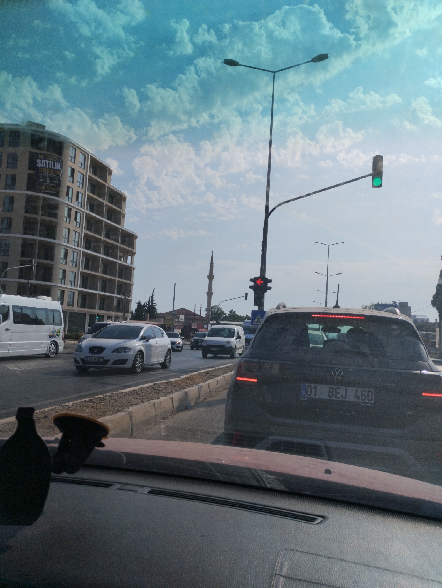 The image depicts a street scene in Erdemli, Turkey, featuring traffic and urban architecture under a partly cloudy sky. The focal point is the rear of a dark-colored Volkswagen T-Cross SUV with the license plate "01 BEJ 460". In front of the SUV, a white SEAT car with license plate "33 BDN 021" is visible, along with a white van and other vehicles. A modern, multi-story building with curved balconies dominates the left side of the frame, displaying a "SATILIK DAİRELER" (For Sale Apartments) sign. A minaret of a mosque is present in the background. Traffic lights are visible, with one showing a green light and another showing a red left arrow. The foreground shows the interior of a vehicle, including a dashboard, a suction cup mount, and what appears to be a bottle. The overall environment suggests a typical urban setting with ongoing traffic flow.