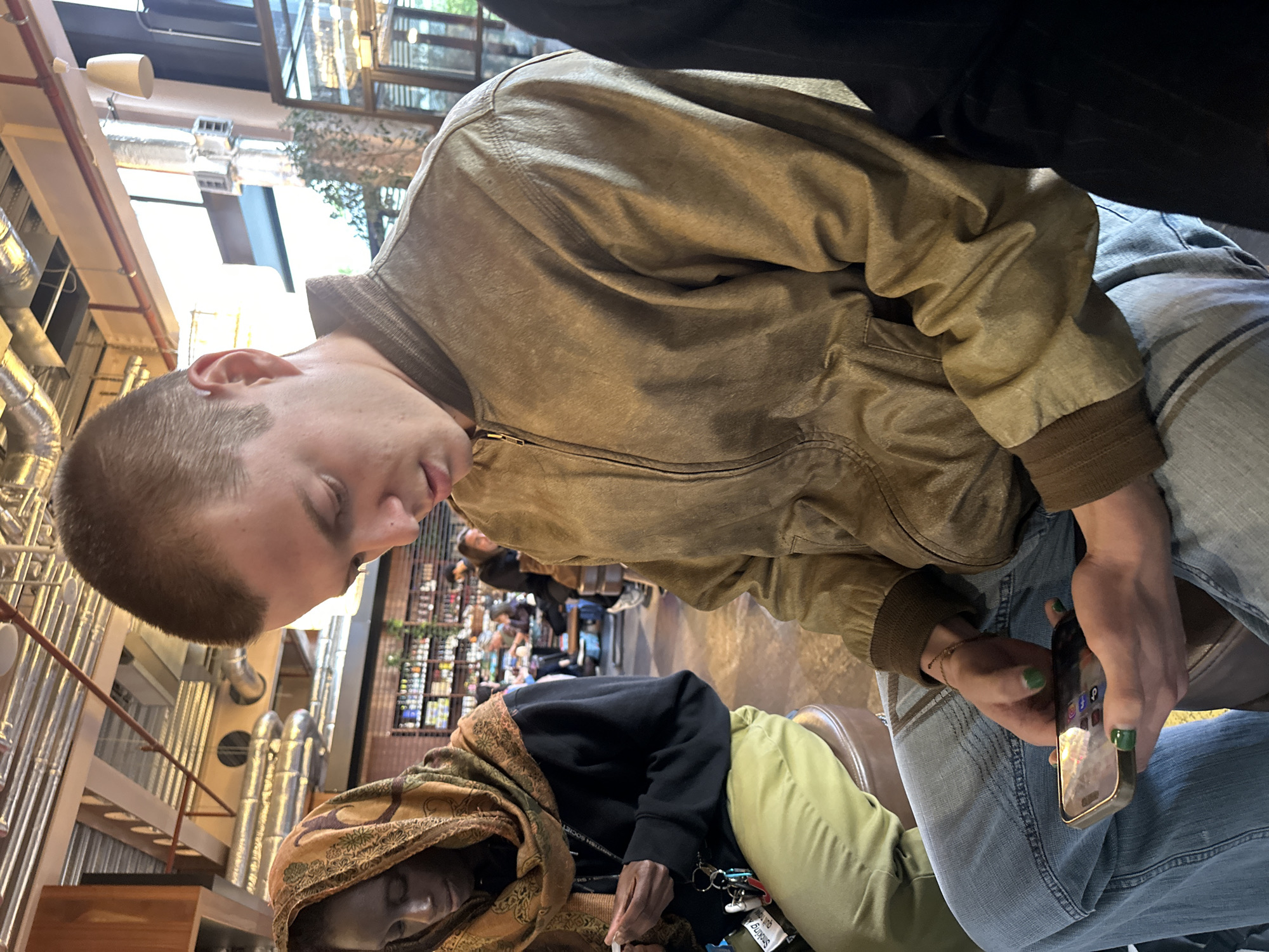 An indoor scene, likely a restaurant or bar in London, UK. A young fair-skinned individual with short hair sits to the right, wearing a tan jacket and jeans, holding a smartphone with green painted fingernails. To the left, another person wearing a patterned headscarf, black hoodie, and light green pants sits. In the background, a bar setup with multiple bottles, and other people are visible. Metallic pipes are seen on the ceiling.