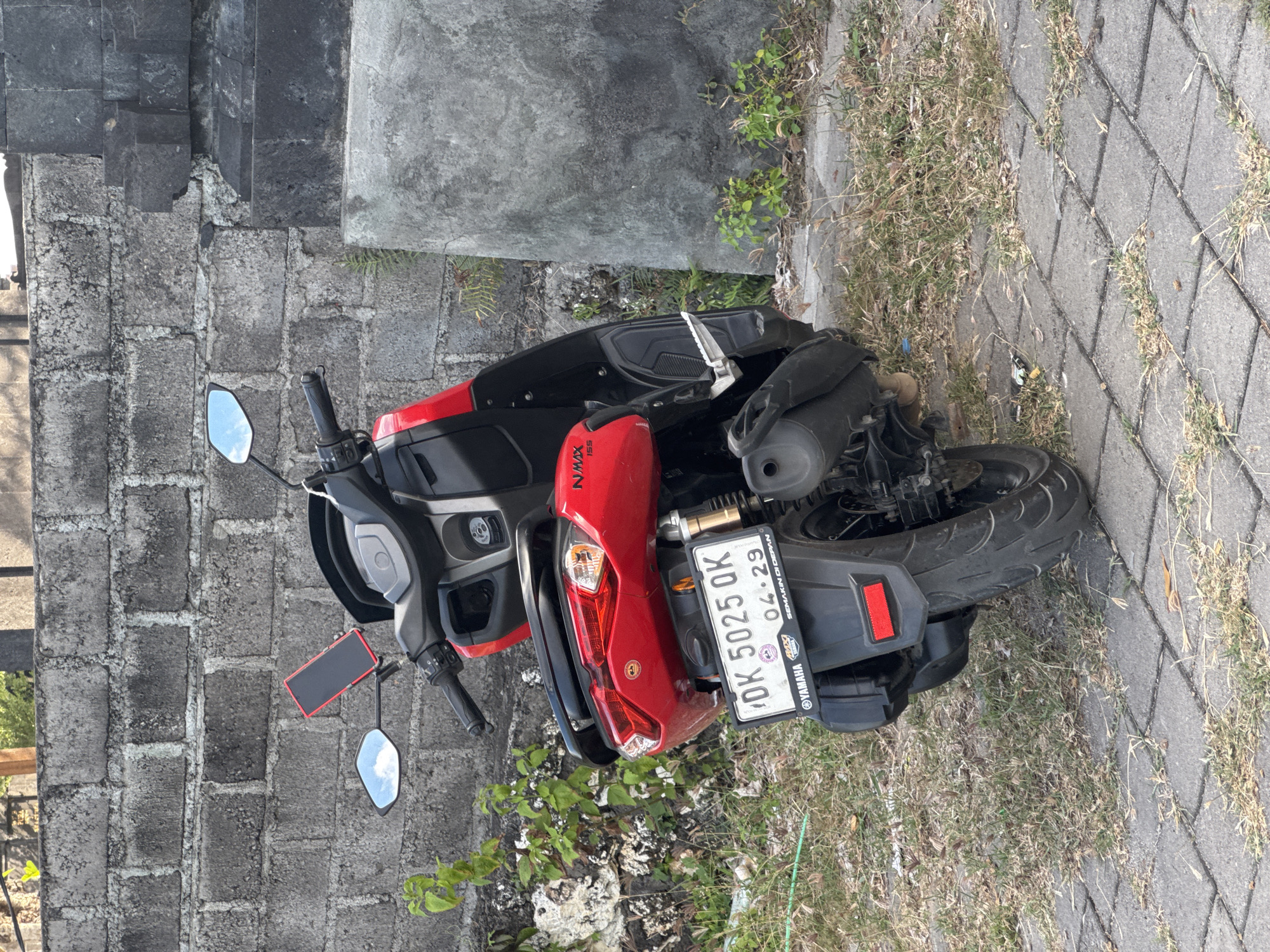 A red and black Yamaha NMAX 155 scooter is the central subject of the image, appearing to be parked or abandoned outdoors. The scooter is positioned on a paved surface made of square concrete blocks, with patches of grass and weeds growing between them and alongside. In the background, there is a rough concrete block wall, typical of local construction.
The scooter features a smartphone mounted on its handlebars, and its rearview mirrors reflect a bright blue sky with some clouds, indicating daytime and clear weather. The scooter itself appears to be in used condition, with some dirt visible.
Notable text includes "NMAX 155" on the side of the scooter and a visible Indonesian license plate that reads "DK 5025 QK" with "04.29" printed below it, likely indicating the month and year of validity or registration. The brand "YAMAHA" is also visible on the mudguard near the license plate. Small, partially obscured text on the license plate frame appears to include "SEMAKAN DENPASAR". The overall scene suggests a typical roadside or outdoor parking area in Kuta Selatan, Indonesia, aligning with the provided location context. No people are visible in the image.