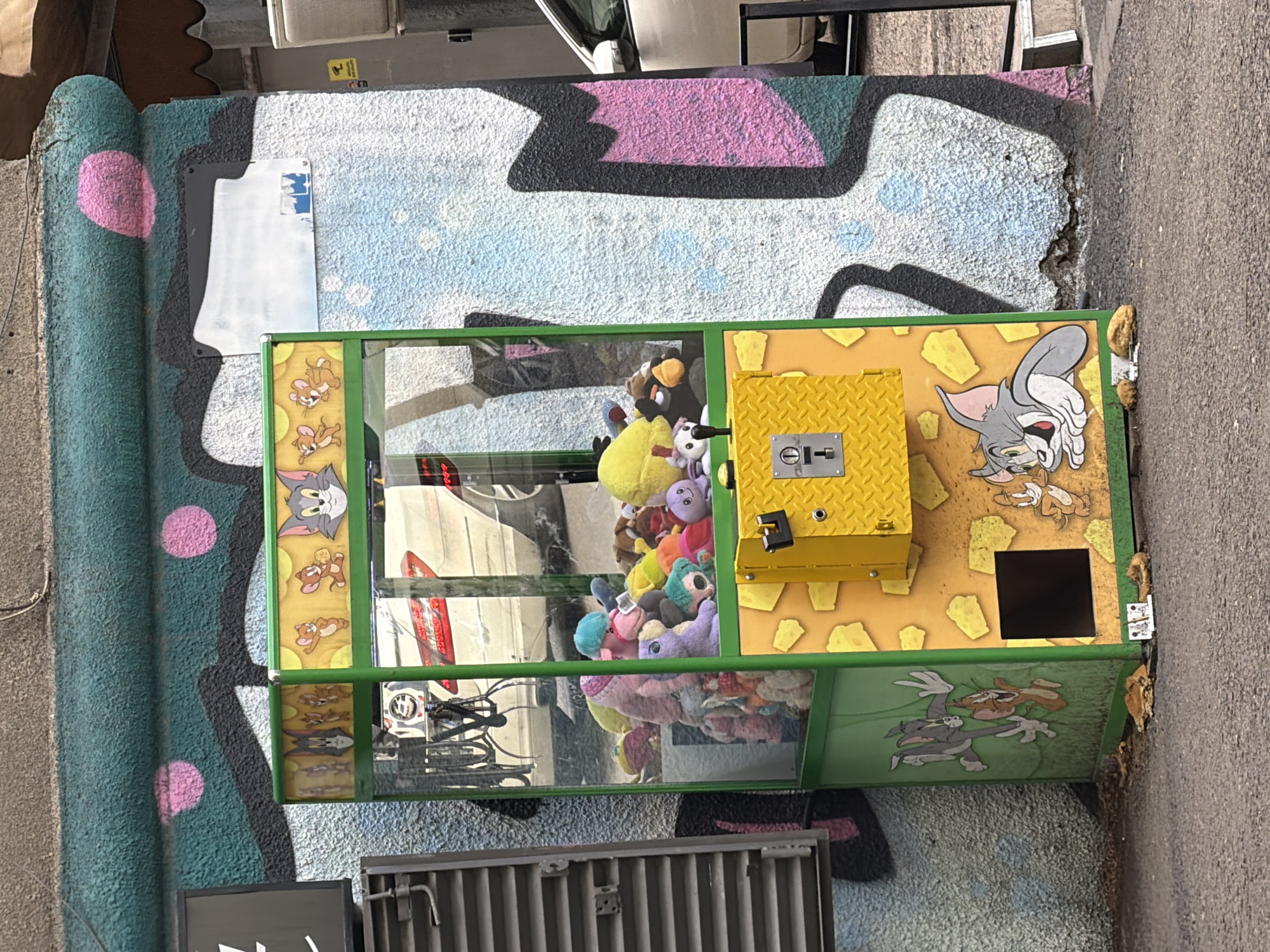 A tilted, eye-level shot depicts a "Tom and Jerry" themed claw machine positioned against a graffiti-laden wall in Tbilisi, Georgia. The claw machine, filled with stuffed animals, features cartoon depictions of Tom and Jerry on its sides. A yellow, diamond-plated coin mechanism is visible. The wall behind the machine is adorned with graffiti, with shades of pink, black, and blue. The ground appears to be asphalt. A vehicle is partially visible in the background.