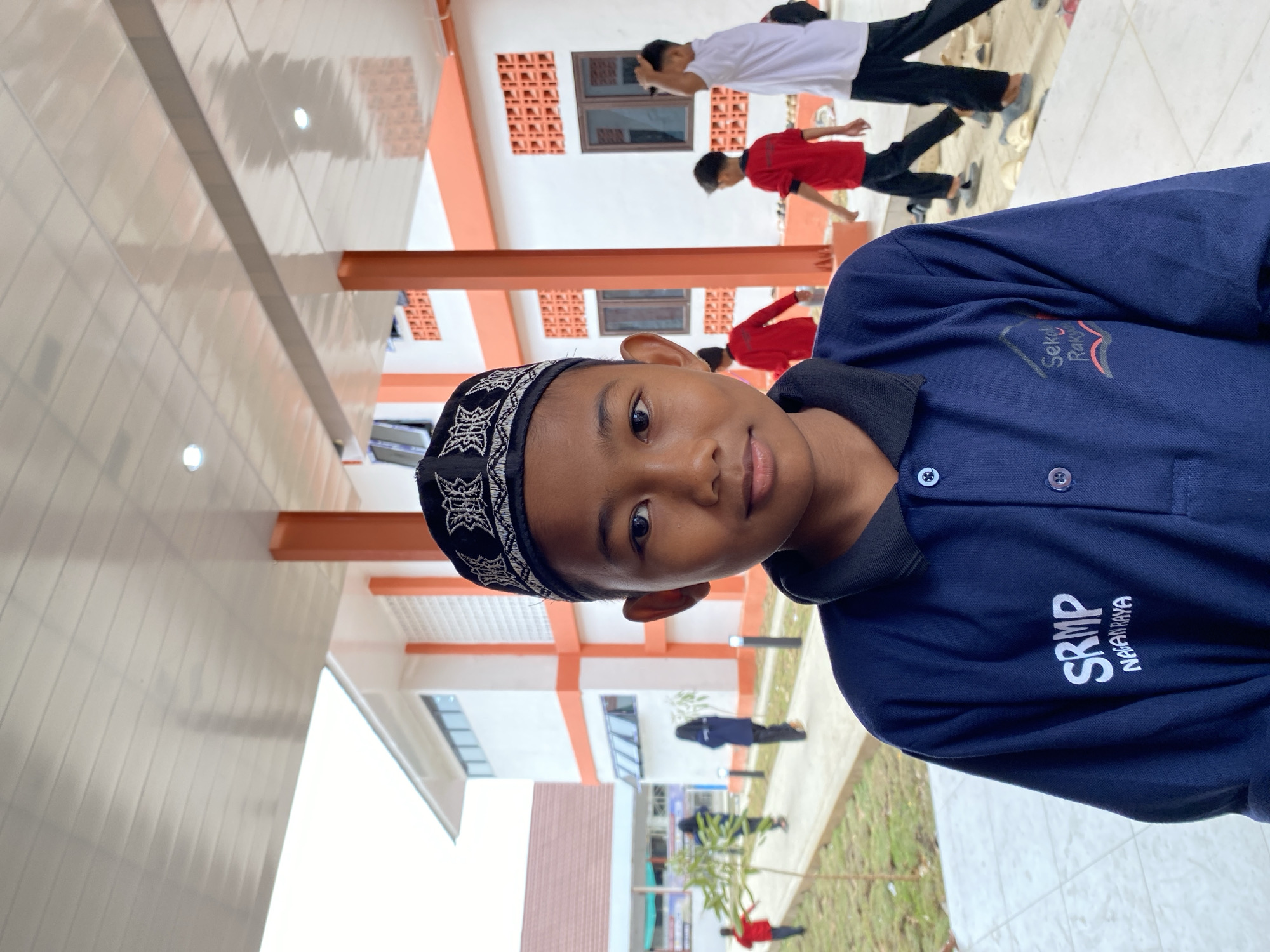 A medium shot captures a young boy wearing a navy blue polo shirt and a black prayer cap, facing forward with a slight smile. The polo shirt has white lettering that reads "SRMP" and "NAGANRAYA" on the left chest, and a logo with text that appears to be "Sekolah Rakyat" on the right. He stands in front of an outdoor walkway with an orange and white building in the background. Several other children, some in red shirts, are visible walking in the distance along the walkway. The environment appears to be a schoolyard or campus, with a tiled floor and green grass to the left. The lighting suggests it is daytime, possibly overcast due to the diffused light. The building features orange pillars and white walls with orange accents, as well as visible windows. The overall scene depicts a casual, everyday moment at what is likely an educational institution.