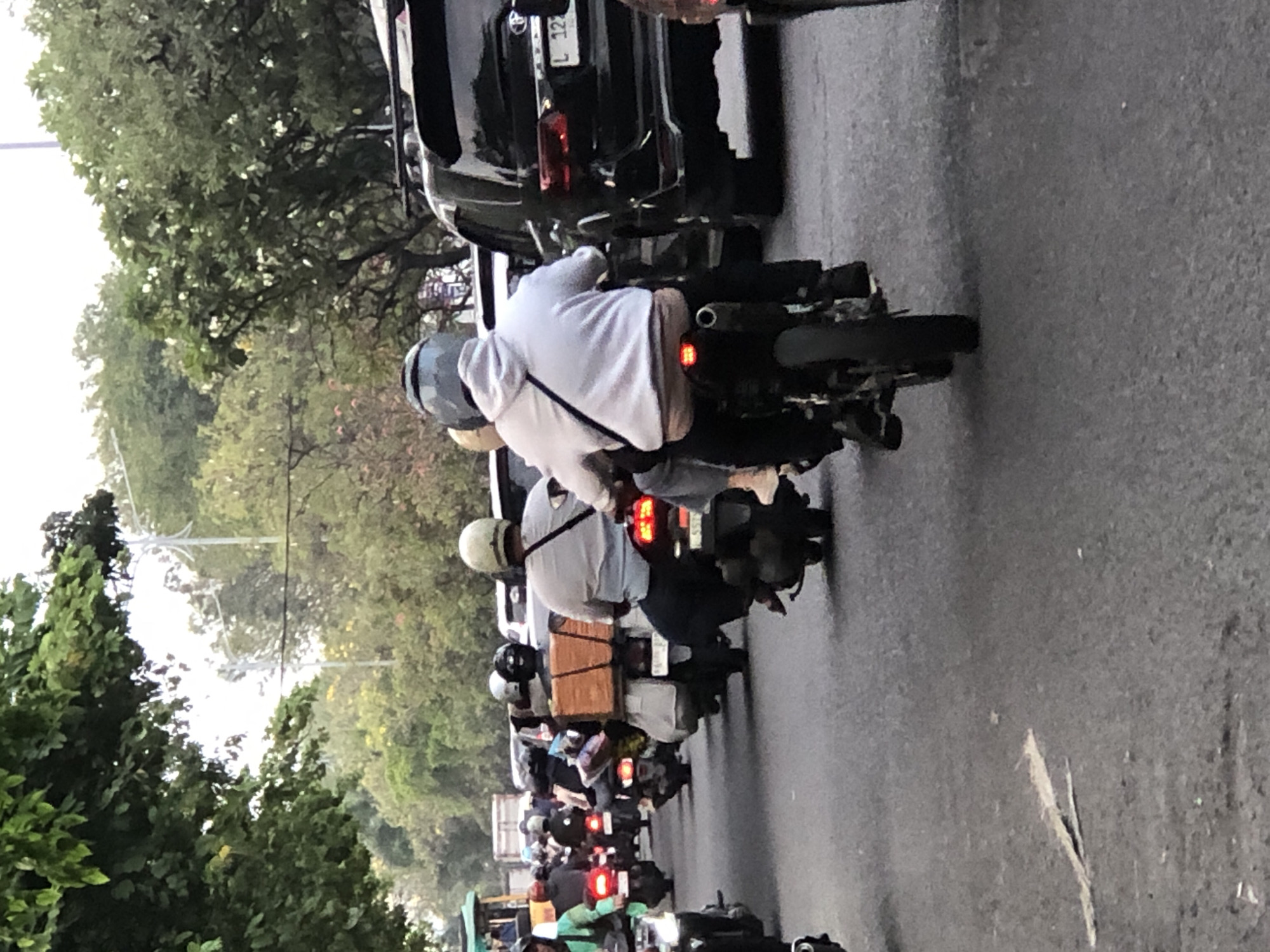 The image depicts a street scene in Sumorame, Indonesia, characterized by heavy traffic congestion involving numerous motorcycles and a few cars. The perspective is from behind a motorcyclist wearing a light-colored hoodie and a helmet, suggesting a journey through the city. Several other motorcycles are visible, some carrying passengers and considerable cargo, such as boxes and bags, indicating the movement of goods or personal belongings. A black SUV is also present in the traffic, highlighting the mix of vehicles. The presence of trees lining the street and a cloudy sky suggests an urban or suburban environment. The overall impression is one of a busy, potentially everyday commute or transportation activity in Sumorame.