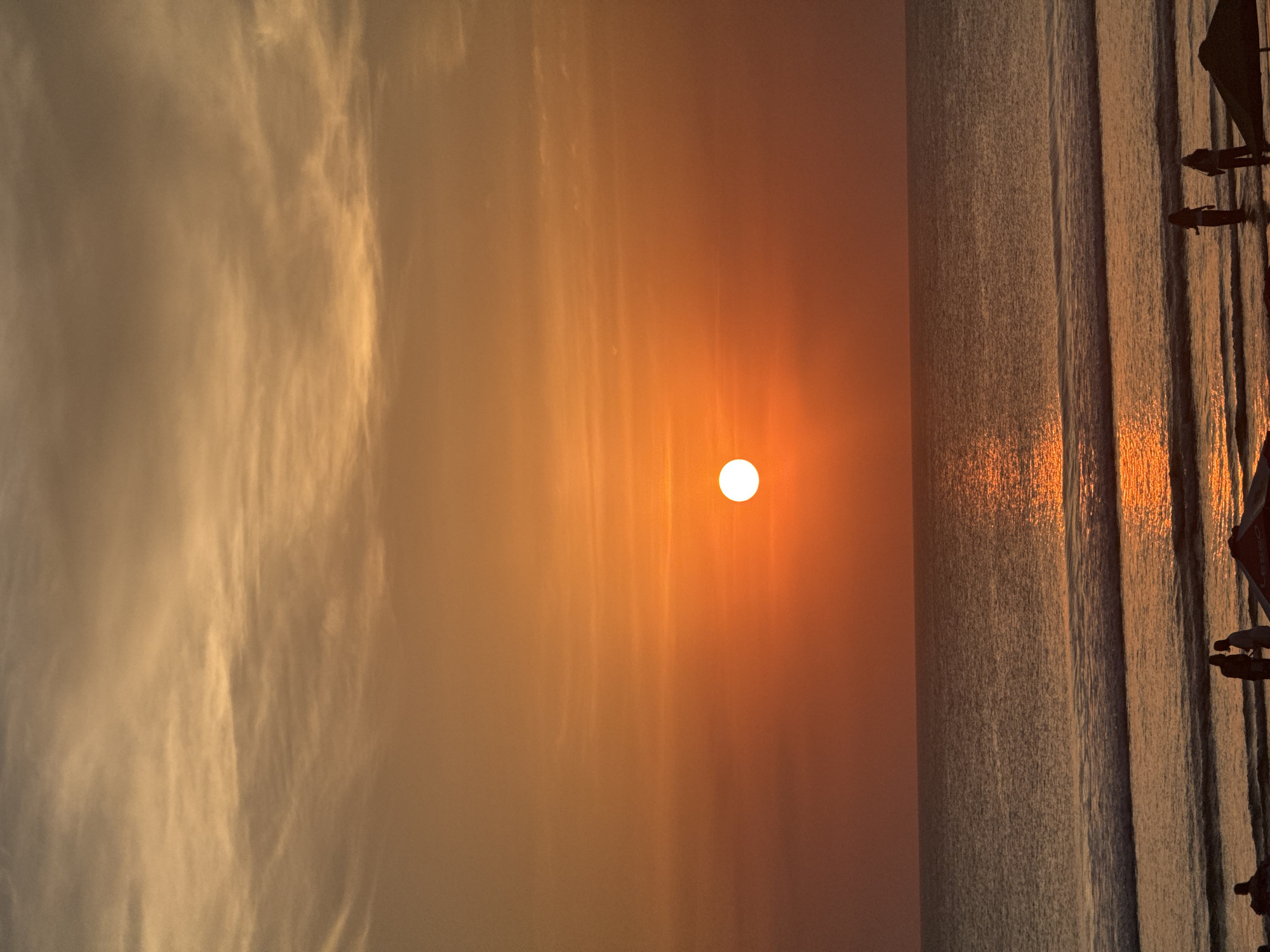 The image depicts a serene sunset scene at Kuta, Indonesia. The sun, a bright, white orb, is positioned in the sky, casting a warm, orange glow across the scene. Wispy clouds streak the sky, tinged with the sunset's colors, suggesting fair weather. The ocean is calm, with gentle waves rolling towards the shore, reflecting the golden light of the setting sun. Silhouettes of people can be seen on the right side of the image, standing near what appears to be a pier or a boardwalk. They are indistinct figures, adding to the tranquil atmosphere. One person is closer to the camera, with their reflection visible in the water, while others are further away. The overall mood is peaceful and picturesque, capturing the beauty of a sunset by the sea. The time of day is clearly sunset. There is no visible text in the image.
