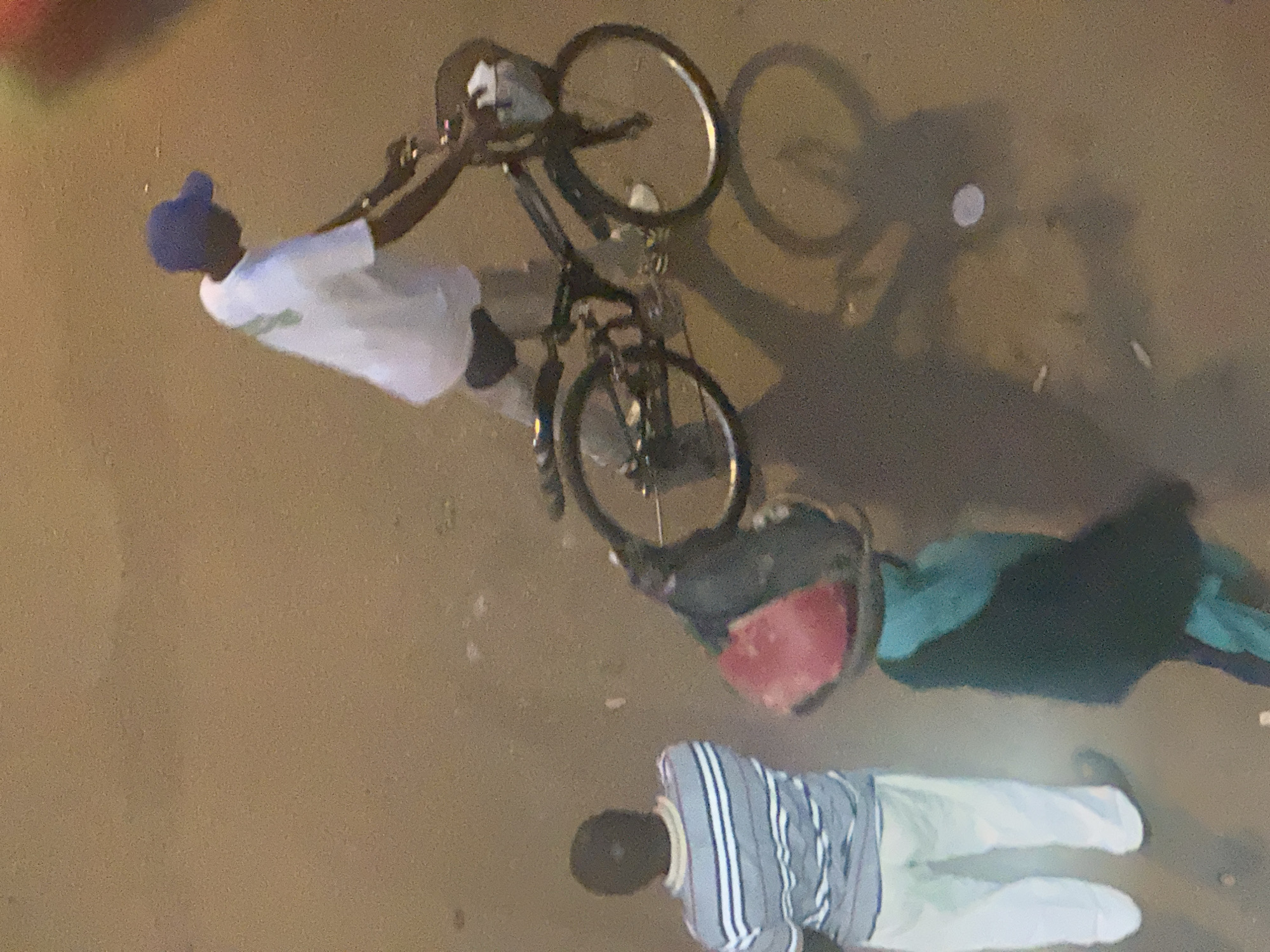The image shows a top-down view of two individuals and a bicycle on a paved surface, likely a street or sidewalk in Bukuru, Nigeria. One person, wearing a white t-shirt and a blue cap, is standing next to a bicycle. The bicycle has a black frame and appears to have a bag attached to it, containing a red object. The person is holding the handlebars of the bicycle. In the foreground, another individual is lying down on the ground, wearing a striped long-sleeved shirt and light-colored pants. This person is facing away from the camera and appears to be resting or possibly in distress. The lighting suggests it is nighttime or dusk, as shadows are prominent. The ground is a neutral, sandy brown color with some debris visible. The overall scene depicts a moment of stillness, possibly after activity, with one person engaged with their bicycle and the other resting on the ground. No visible text is present in the image.