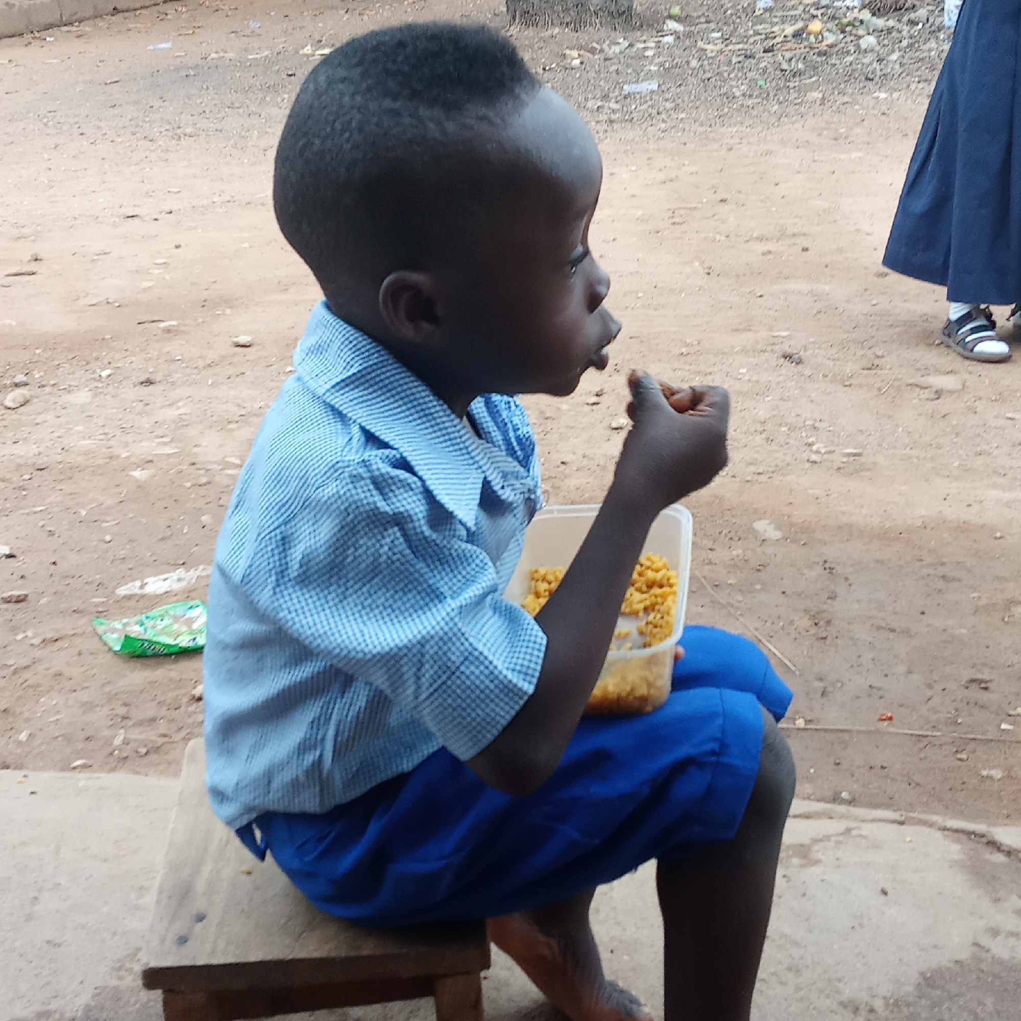 A young boy is sitting on a wooden stool, holding a plastic container filled with cooked corn and eating some. He is wearing a blue checked shirt and blue shorts, likely a school uniform. The location appears to be outdoors, possibly in Akwanga, Nigeria, given the context. The ground is dirt and concrete, with some litter visible. In the background, part of another person wearing a blue skirt and white socks with sandals can be seen. The setting suggests a relatively poor or underdeveloped area.