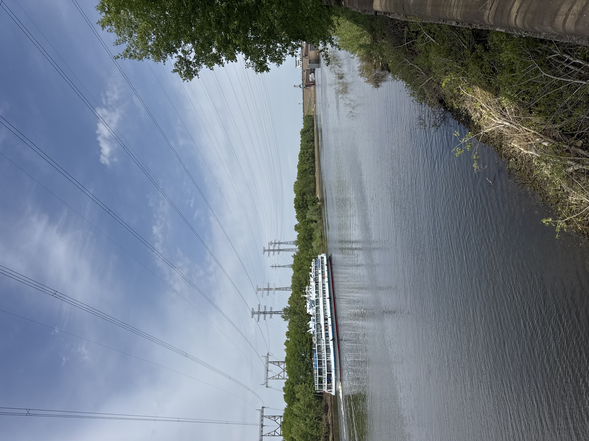 The image depicts a river scene in Chaikovsky, Russia. A multi-deck passenger ship is traveling on the river. Overhead power lines stretch across the sky along the riverbank. The banks are lined with trees and vegetation. A concrete structure is visible along the right riverbank. The sky is overcast with some visible clouds.