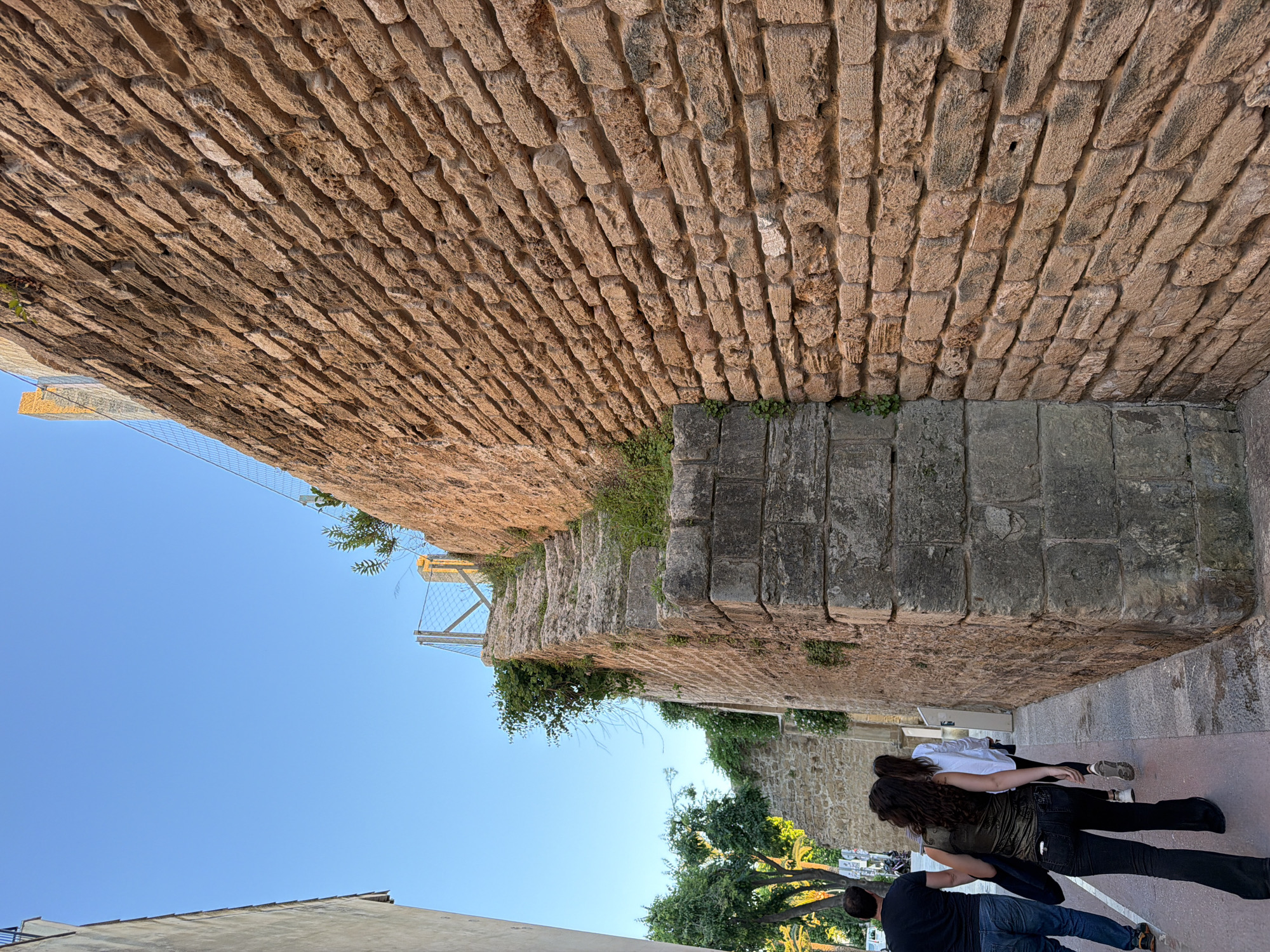 The image captures a view within the city of Alcúdia, Spain. The scene is dominated by the presence of old stone walls, likely part of the city's historical fortifications. The walls are constructed of large, tan and brown textured stone blocks. There is some green vegetation growing out of the cracks and crevices of the wall.
A walkway is visible between the walls, on which three people are standing. Two women, one wearing a white shirt and the other wearing a green shirt and black pants, are standing close together. A man, wearing a black shirt and blue jeans, is standing close to them.
In the upper-left corner of the image, there is a glimpse of a blue sky and a metal structure that appears to be part of a walkway or bridge attached to the stone wall. Below, other buildings are visible. A tree with yellow flowers is situated near the bottom right corner of the image.