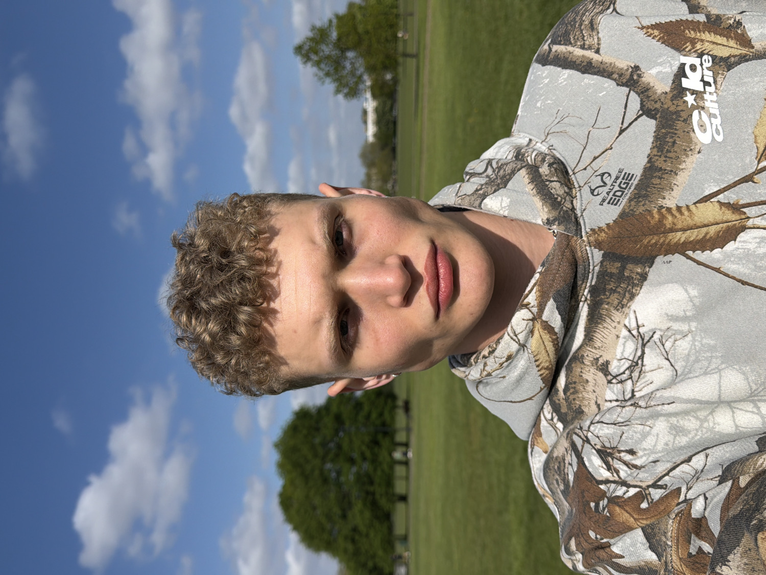 A close-up, eye-level portrait captures a young individual, likely male, looking directly at the camera. He has short, curly light brown hair and a neutral to slightly pensive expression. The person is wearing a hooded sweatshirt featuring a detailed camouflage pattern of white, grey, and brown branches and leaves. Visible logos on the sweatshirt include "REALTR EE EDGE" on the left chest and "Guld culture" near the right shoulder/hood.
The setting is an outdoor park or large grassy field, consistent with locations found in London, United Kingdom. Behind the subject, a vast expanse of bright green grass is visible, with distant trees dotting the landscape. The sky is a clear, vibrant blue with scattered white cumulus clouds, indicating a sunny and bright day, likely morning or early afternoon. The natural light illuminates the subject's face. The individual appears to be posing for a photograph, possibly a selfie.