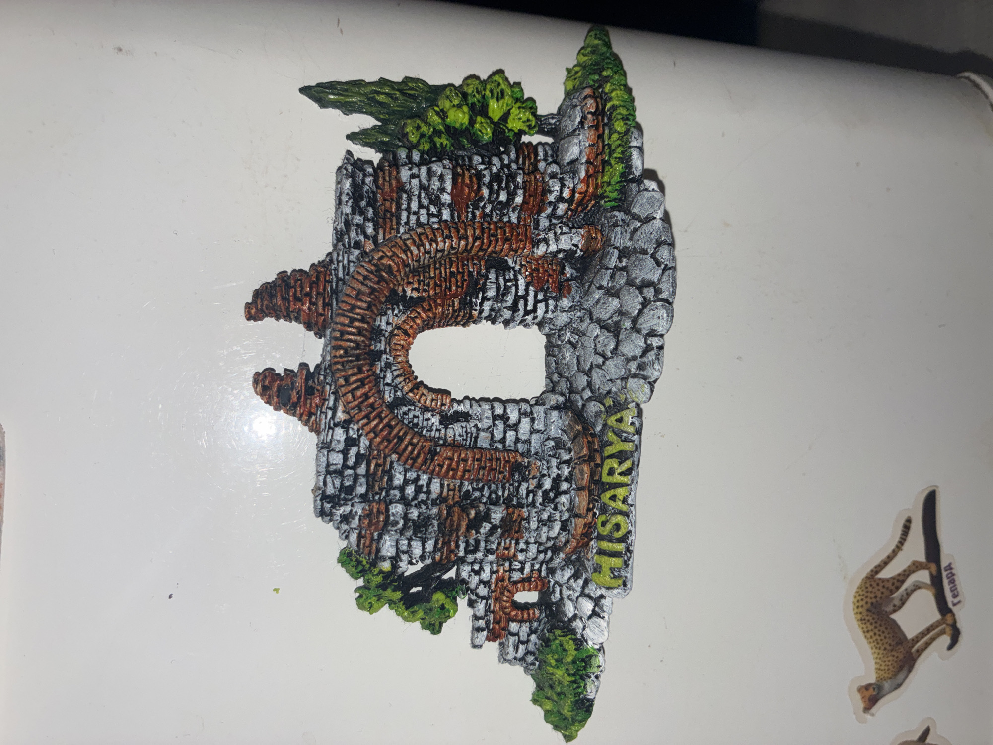 The image shows a magnet on a white surface, possibly a refrigerator. The magnet is a three-dimensional representation of ancient ruins with an arched gateway. The ruins are depicted with gray stonework and red brick accents, overgrown with green foliage and trees. The word "HISARYA" is written in yellow letters on the stone base of the ruins. Below the ruins, there is a sticker of a cheetah with the word "Гепард" written on it in Cyrillic.