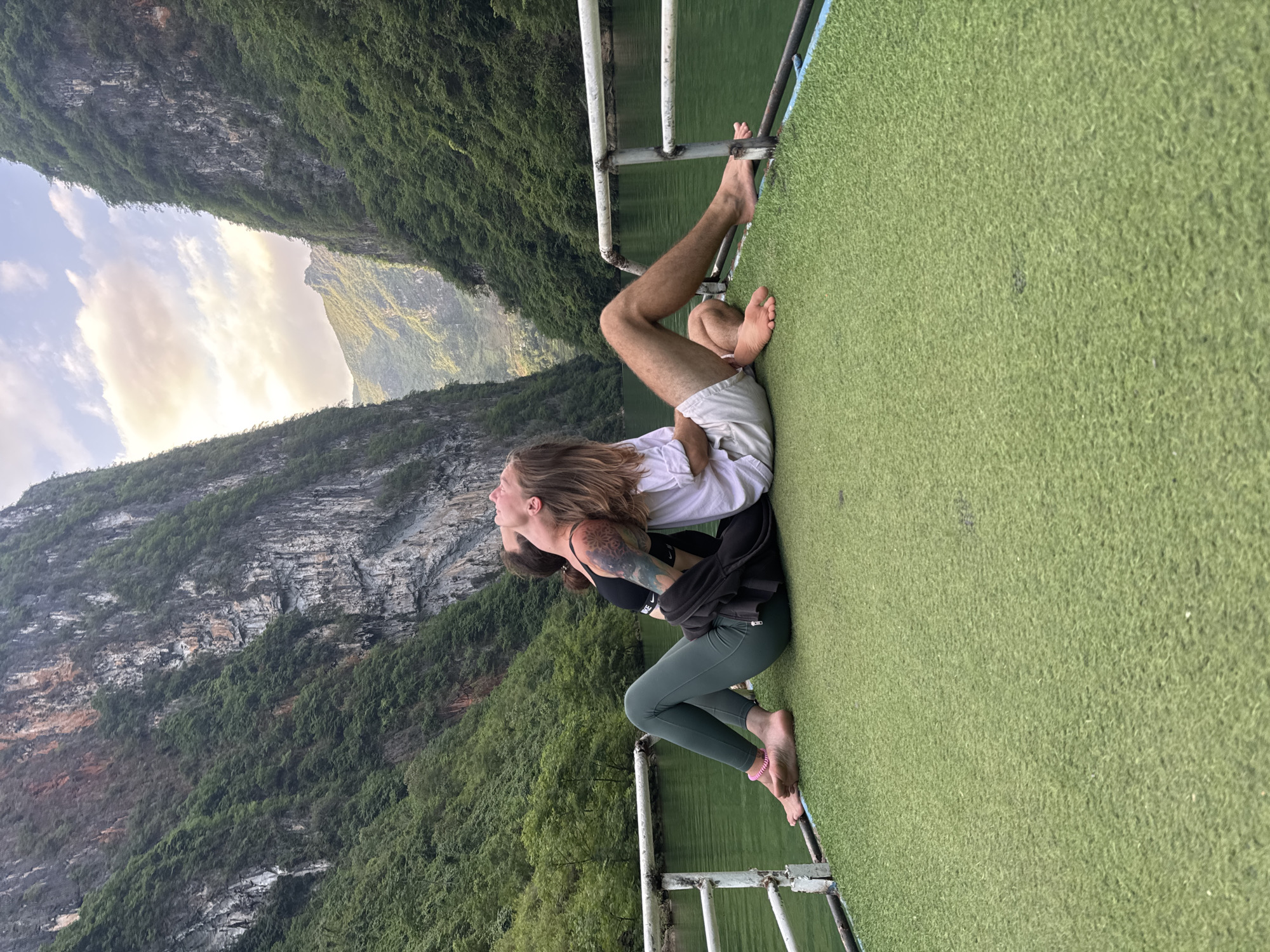 A medium shot depicts a couple in an embrace, positioned on the deck of a boat, with a mountainous backdrop. The man has long blonde hair and wears a white shirt and shorts. He is cradling the woman, who sports a black sports bra, green leggings, and a black jacket. Both are barefoot, with their feet near the edge of the boat deck, which is covered in green artificial turf. The boat's railing is visible behind them. The landscape reveals rugged, tree-covered mountains and a sky with clouds illuminated by sunlight. This scene is in Đồng Văn Commune, Vietnam.