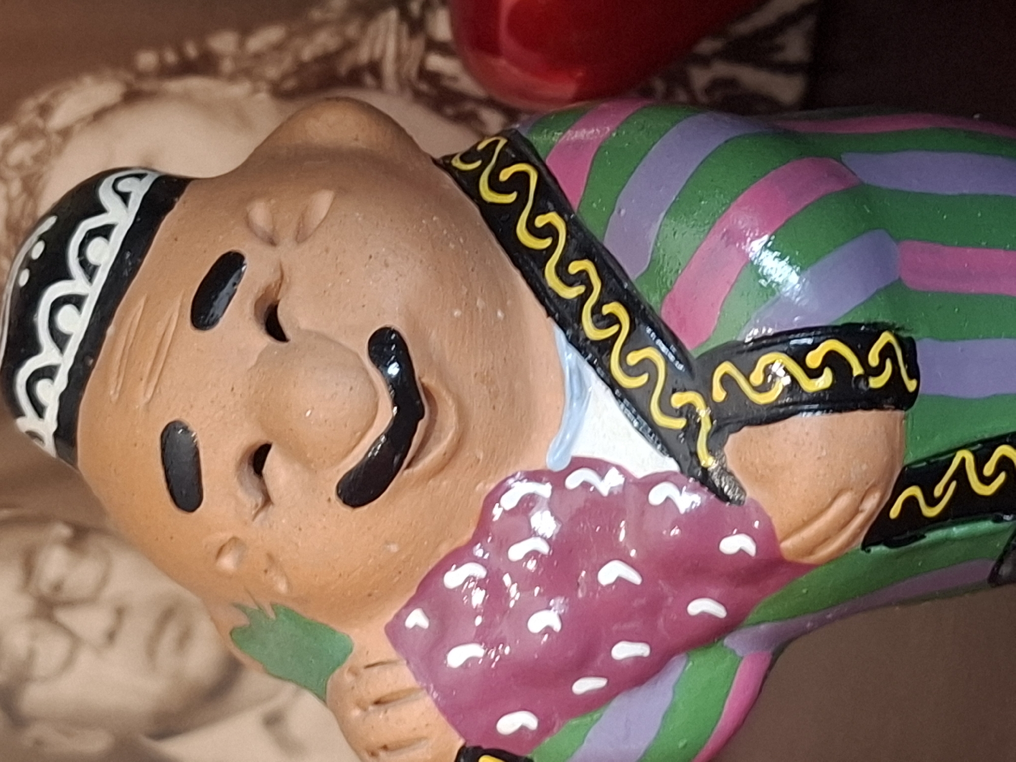 The image shows a close-up of a ceramic figurine of a man. The figurine is smiling and wearing a patterned hat and a colorful striped robe with a black and yellow trim. In the background, there is a faded black and white photograph of a man wearing glasses, and to the right, a blurred red object, possibly an apple, is visible. The figurine's skin is a light brown color, and its eyes and mustache are black. The robe features alternating stripes of green, purple, and pink. The hat is black with a white decorative pattern. The overall scene is a still life, with no apparent actions or interactions taking place. The location of this figurine in Almaty, Kazakhstan is not specified in the image.
