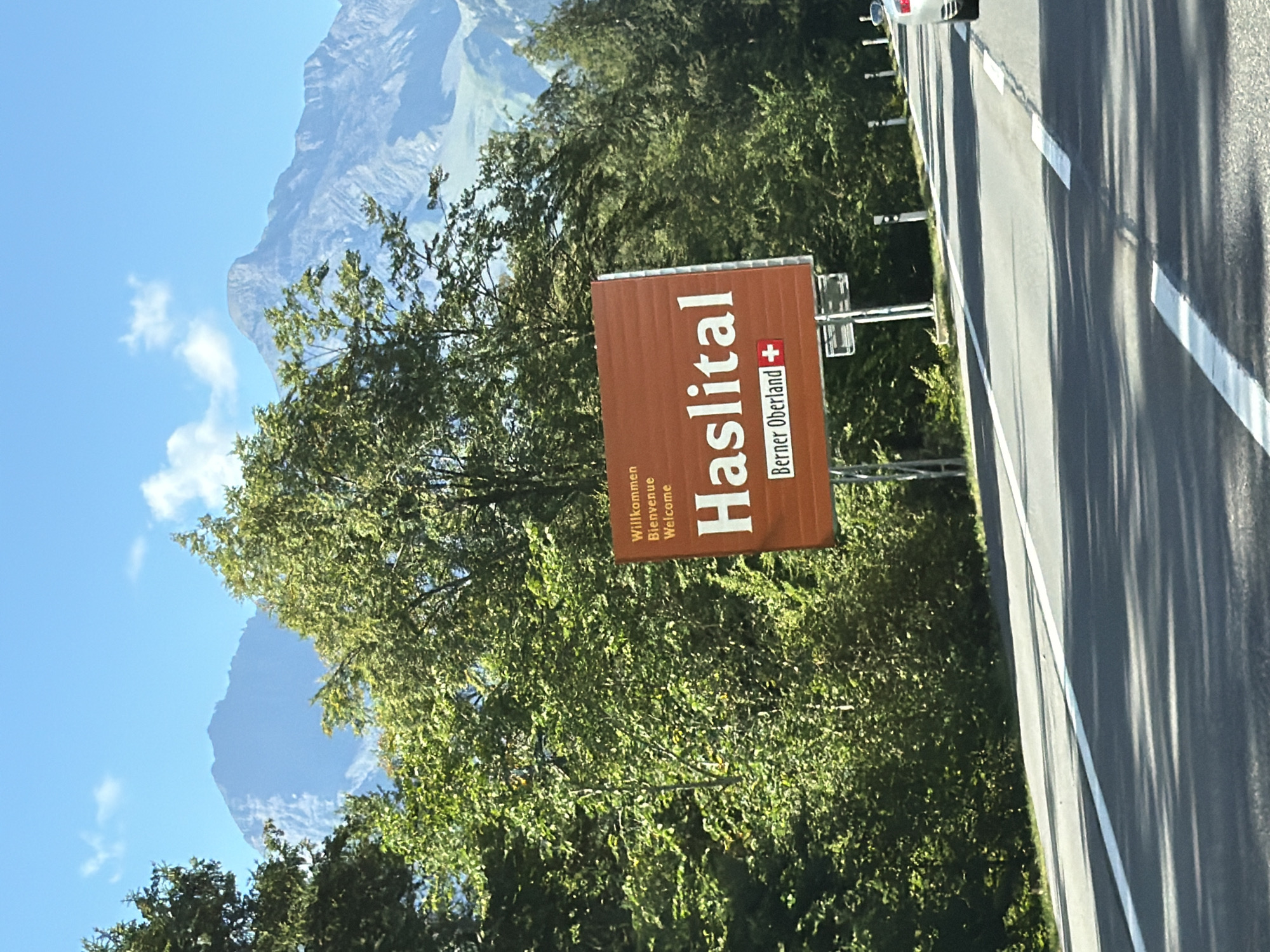 A roadside sign welcomes visitors to Haslital and the Bernese Oberland, with the Swiss flag on the right. The sign is a warm, reddish-brown color and features white lettering. Above the main name, it says "Willkommen Bienvenue Welcome" in smaller text. The sign is framed by lush green trees and a backdrop of majestic, hazy mountains under a clear blue sky dotted with a few white clouds. A paved road with white lane markings stretches out in front of the sign, suggesting a scenic driving route. The perspective is from a vehicle traveling on the road, indicated by the slight tilt of the sign and the road ahead.