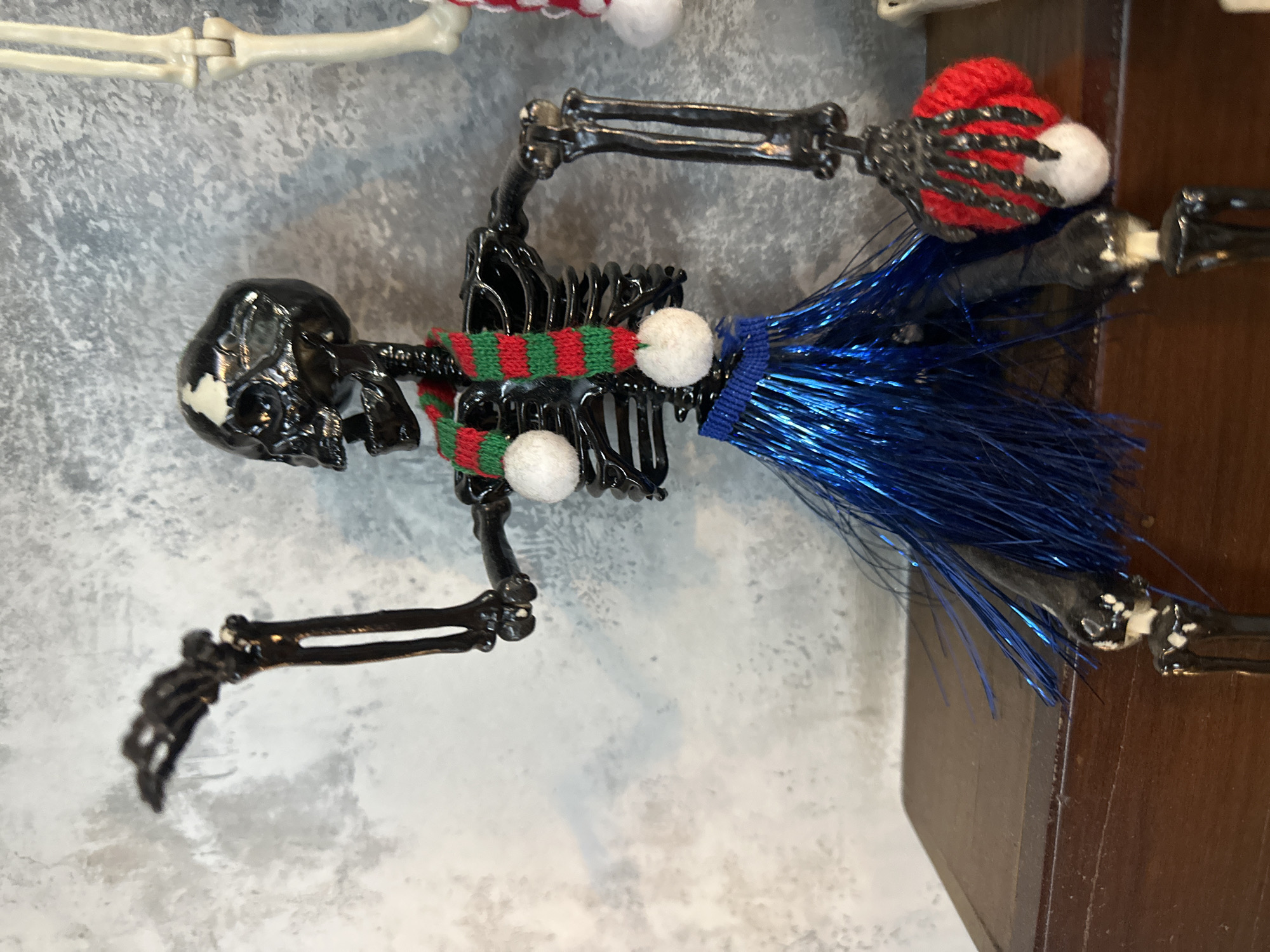 A black plastic skeleton decorated for Christmas sits on a dark wooden surface in front of a textured grey wall. The skeleton is wearing a red and green striped scarf with white pom-poms, and a shiny blue tinsel skirt. It is holding a white pom-pom with a red knitted cap in its right hand. The skeleton's left arm is raised in the air. The scene is not located in Đà Nẵng, Vietnam.