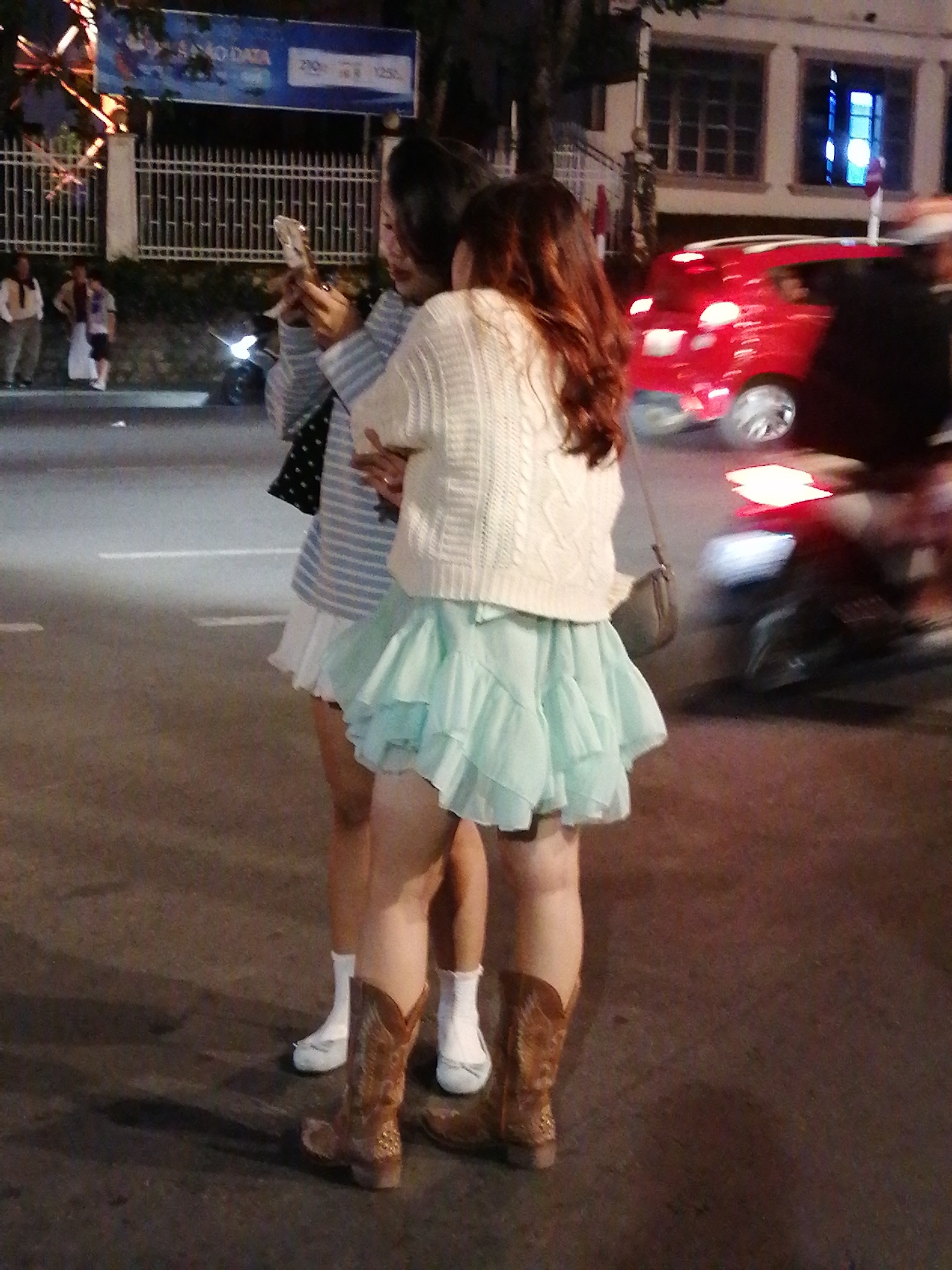 Two young women are standing on a paved street at night, possibly in Da Lat, Vietnam. The woman on the left is wearing a striped shirt and a white ruffled skirt. The woman on the right is wearing a white knitted sweater and a light blue ruffled skirt. Both women have their hair down. The woman on the left is holding a cell phone. The woman on the right has a small purse slung over her shoulder. In the background, there are buildings with illuminated windows, a white fence, and a red car driving past. There are also street lights visible. The ground is a dark asphalt surface.