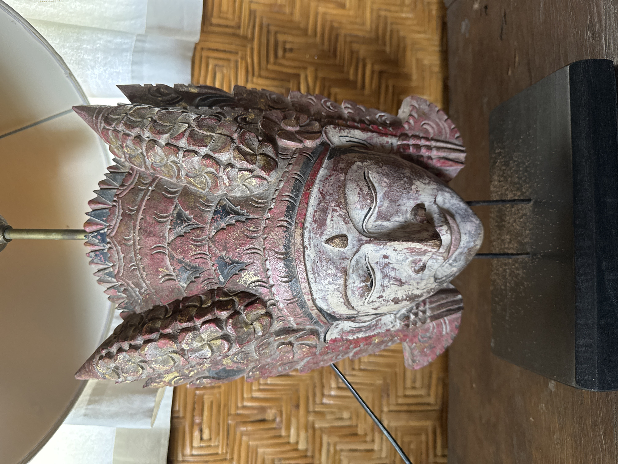 The image features a intricately carved wooden head displayed in an indoor setting. The head, possibly depicting a Buddha or a traditional deity, has a serene expression with closed eyes and a gentle smile. It is adorned with a detailed headdress or crown, featuring layered, scale-like patterns and pointed elements. The carving is painted with a distressed finish, showcasing faded red, white, and black pigments, along with traces of gold that highlight its textured surface.
The carved head is mounted on a slender black metal rod, which is secured into a sturdy, rectangular black display base. To the left, a portion of a lamp with a white, translucent fabric shade is visible, casting a soft light. The background behind the carved head consists of a light brown, woven material, possibly rattan or bamboo matting, forming a distinct geometric pattern. A darker wooden surface, appearing to be a table or floor, is present beneath and to the right of the display.
No activity or event is occurring; the image is a static depiction of an art piece. There is no visible text. The craftsmanship and style of the carving, combined with the woven background, are characteristic of the cultural artifacts and decorative elements found in the Ubud District, Indonesia.