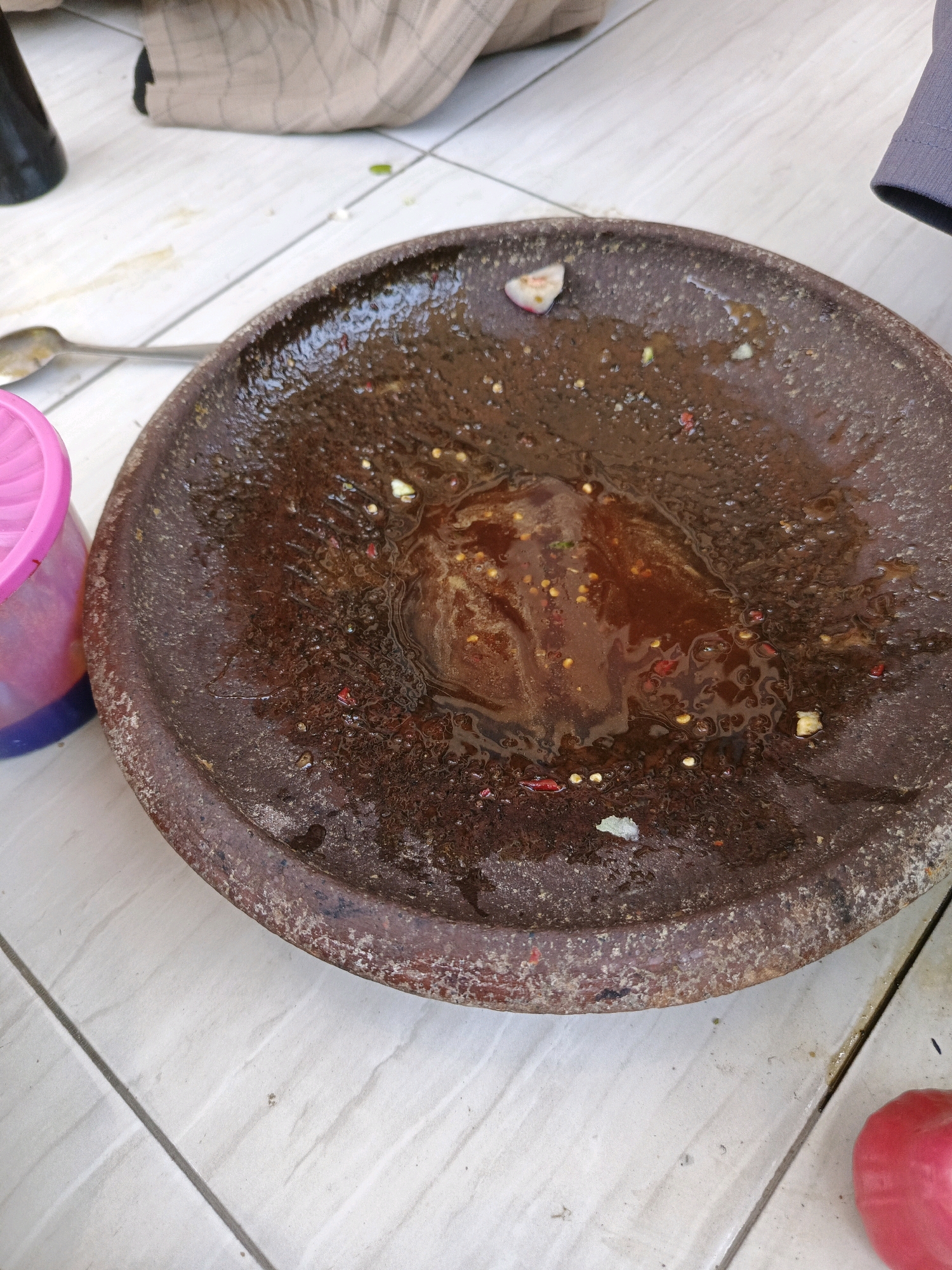 The image shows a close-up view of a mortar and pestle on a tiled floor. The mortar is made of dark brown, rough material and contains a reddish-brown, liquid substance with visible pieces of chili and what appear to be spices. The pestle is not visible. To the left of the mortar, a pink plastic lid of a container is visible, and further to the left, a silver spoon is present. In the background, a portion of a patterned cloth and a dark bottle are visible, suggesting an outdoor or casual setting. The tiled floor is light-colored with visible grout lines. A pinkish-red fruit, possibly a rose apple, is located in the bottom right corner of the image. The overall scene appears to be a food preparation area, likely in a home or a market. The location is specified as Sumbersari, Indonesia.