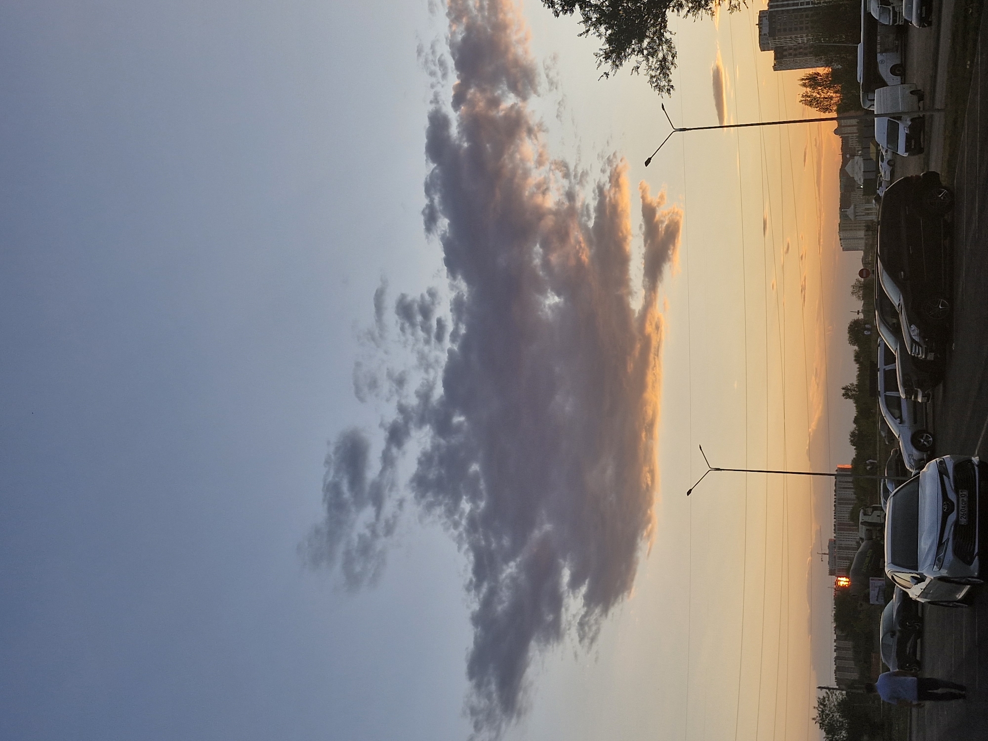 The image captures a dusky sky over a city, with the setting sun casting a warm orange glow across the horizon. In the foreground, a parking lot is filled with various vehicles, including sedans, SUVs, and vans. A single person walks away from the camera across the asphalt. In the midground, streetlights stand tall, and power lines crisscross the sky. The background features a cityscape with apartment buildings and construction cranes, indicative of Astana, Kazakhstan. The clouds overhead are a mix of grays and soft oranges, reflecting the colors of the sunset. The overall scene suggests the end of a day in an urban environment.