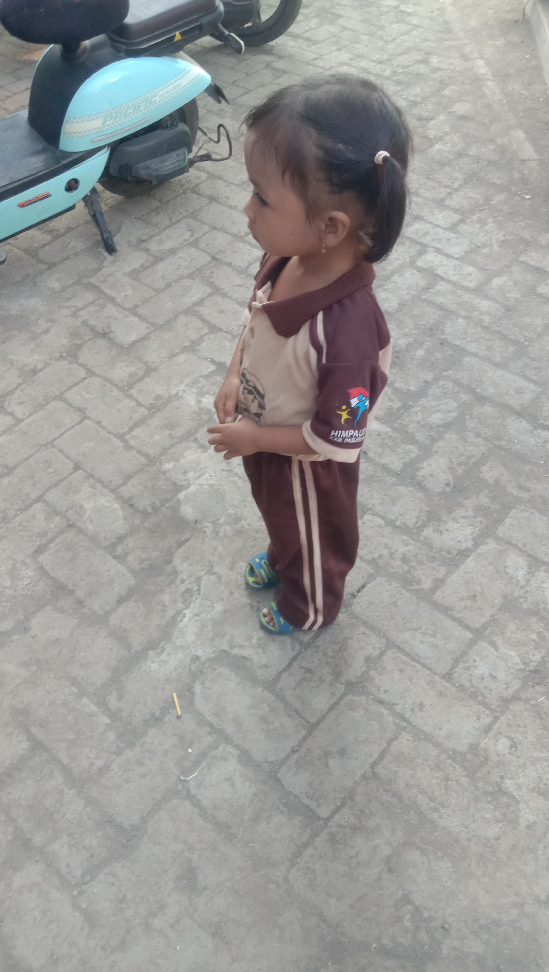 A young child stands on a paved walkway near a light blue scooter in Karang Anyar, Indonesia. The child wears a tan and maroon uniform with a logo on the sleeve and blue sandals. The scooter is parked and appears to be an electric model. The walkway is made of interlocking brick pavers. The lighting suggests it is daytime, possibly late afternoon or early morning given the soft shadows. The background is slightly out of focus, showing more of the paved area and a hint of greenery.