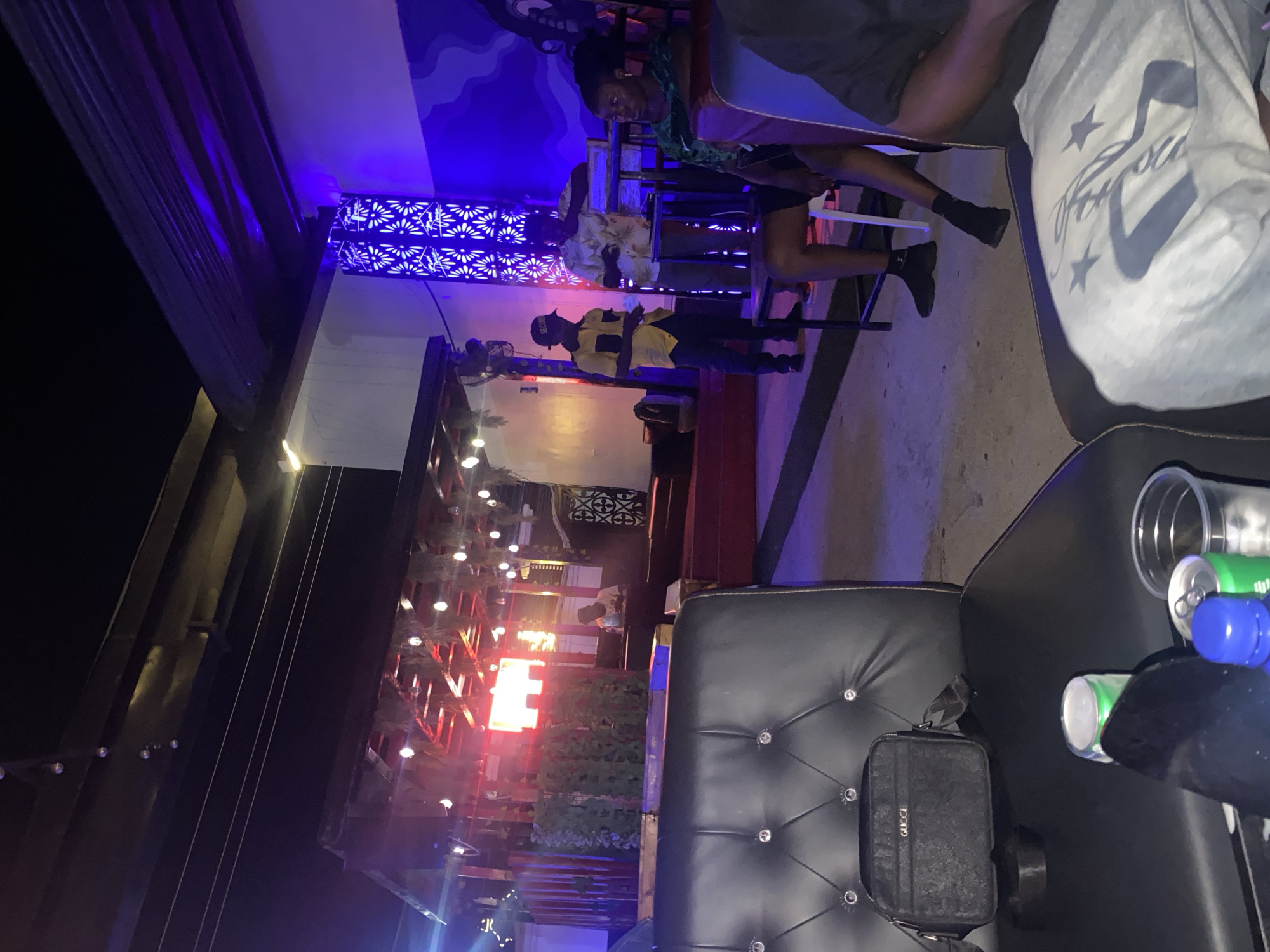 The image depicts an outdoor night scene at a venue that appears to be a bar or club in Howayi, Nigeria. In the foreground, a black leather couch with diamond-like studs is visible. On the couch, a black GUCCI bag is placed next to a green and white soda can, a blue-capped bottle, and a clear plastic cup. Further back, a man in a yellow and black shirt and a black cap is standing near a table. He appears to be talking to another man wearing a patterned shirt. A woman in a green top is seated nearby. The background features an area with red lighting and hanging decorations, possibly a stage or bar area, and a wall with blue decorative lighting. The overall ambiance suggests a lively social gathering.