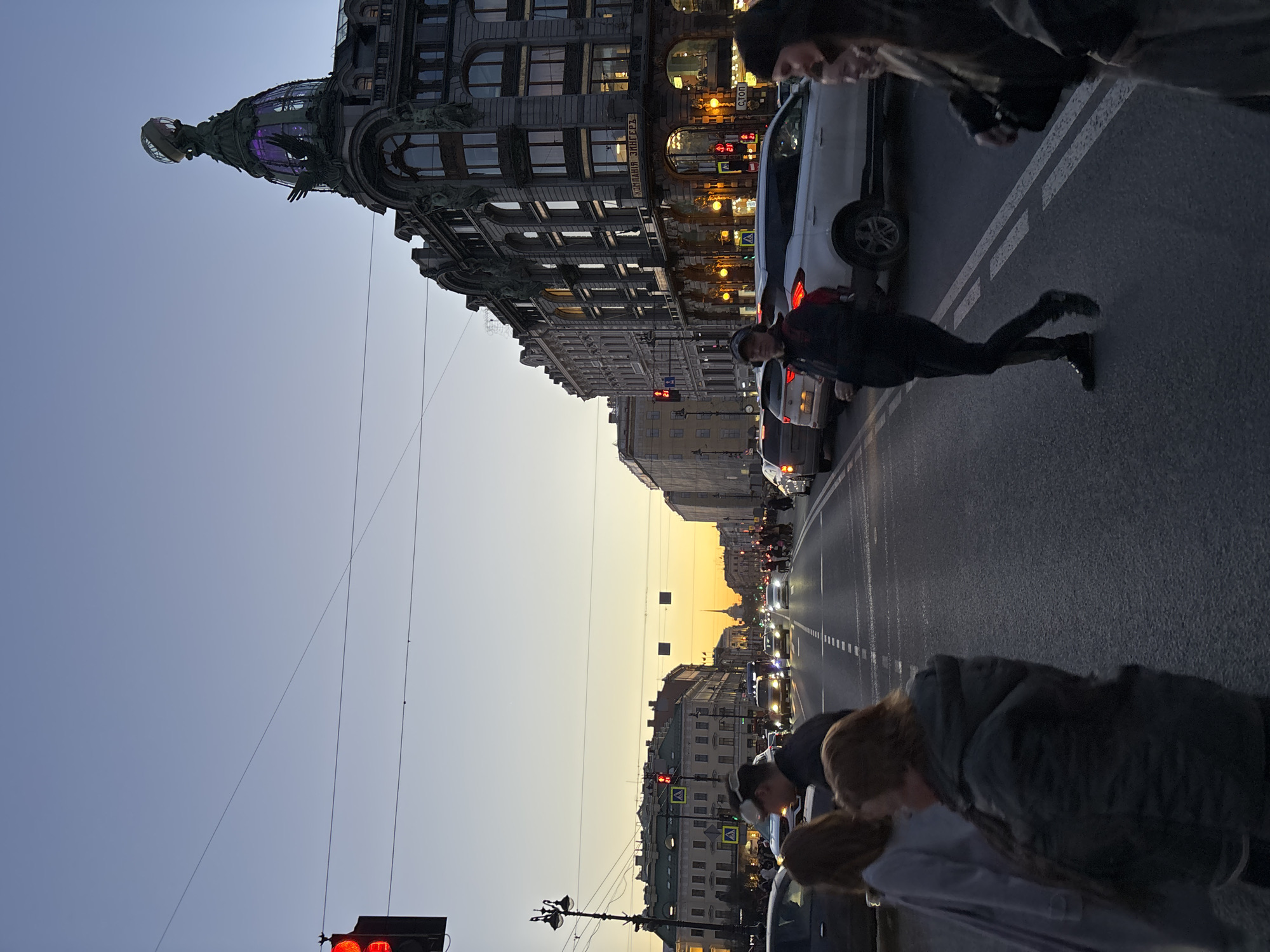 The image depicts a street scene in Saint Petersburg, Russia, at dusk. The prominent architectural feature on the left is the Singer House, a notable building with a distinctive dome. A pedestrian is mid-stride, crossing the street in front of stopped cars, potentially against a red traffic light visible in the foreground. Other people are standing on the sidewalk, observing. Street lights illuminate the scene. The sky is a gradient of blue to orange, suggesting the time of day.