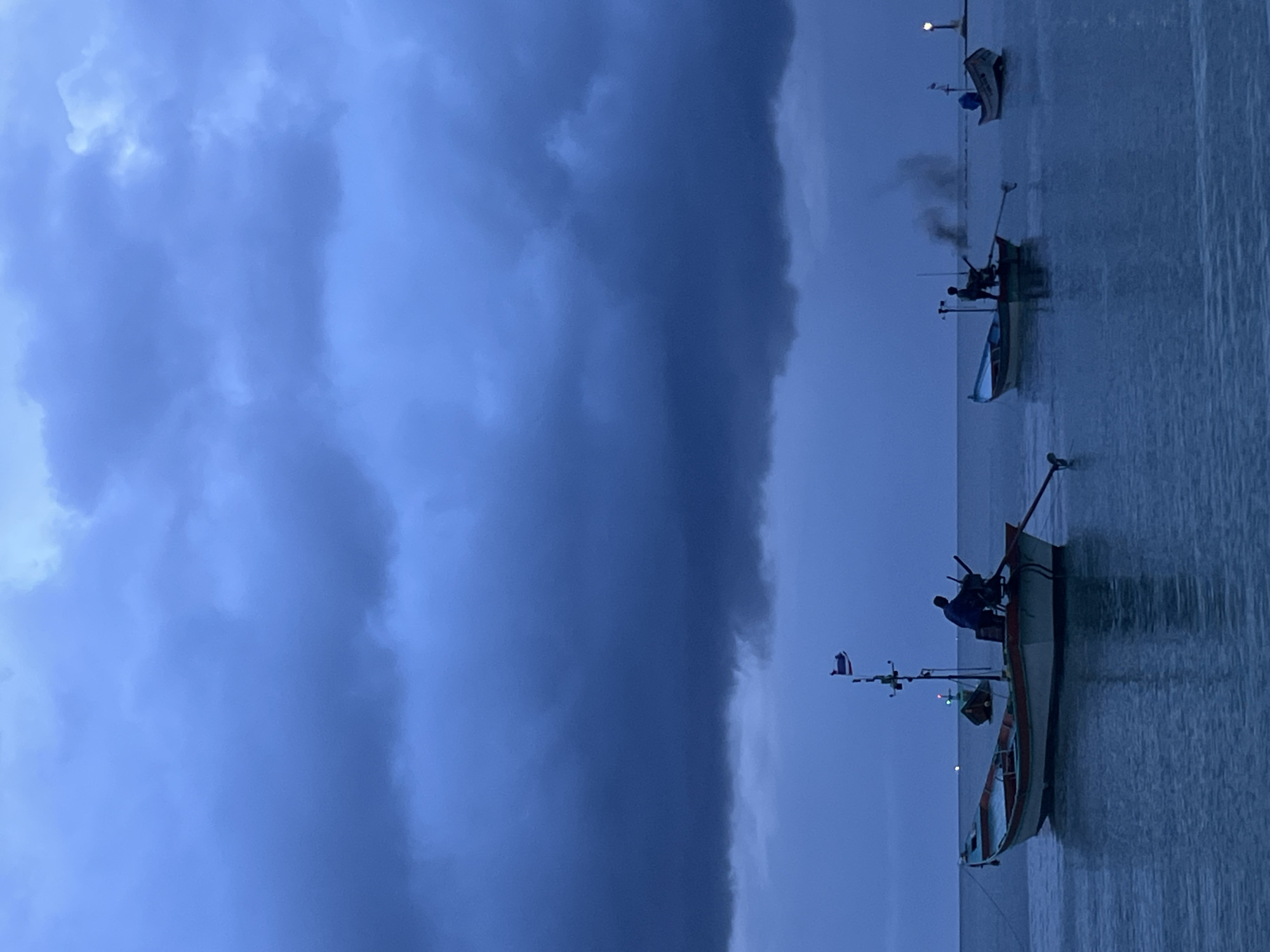 The image depicts a serene yet somber scene of several fishing boats on the water near Ko Pha-ngan District, Thailand, under a heavy, dark sky filled with storm clouds. The water is calm, reflecting the ominous sky above.
In the foreground, a long, narrow fishing boat is anchored. A lone figure is standing in the boat, silhouetted against the dim light, appearing to be engaged in work or observation. The boat itself is painted in muted colors, showing signs of wear and tear. A flagpole with a flag is mounted on the boat.
Further out on the water, two other fishing boats are visible. One of these boats has a figure standing, and smoke is emanating from it, suggesting an engine is running. The other boat appears smaller and further away, with a light visible on its stern, possibly a navigation marker or a small beacon.
The overall atmosphere is one of impending weather change, with the dark clouds dominating the upper half of the frame. The lighting is diffused and dim, indicating either early morning or late evening. The scene conveys a sense of quiet anticipation before a storm, with the fishing boats being the sole subjects against the vast expanse of water and sky.