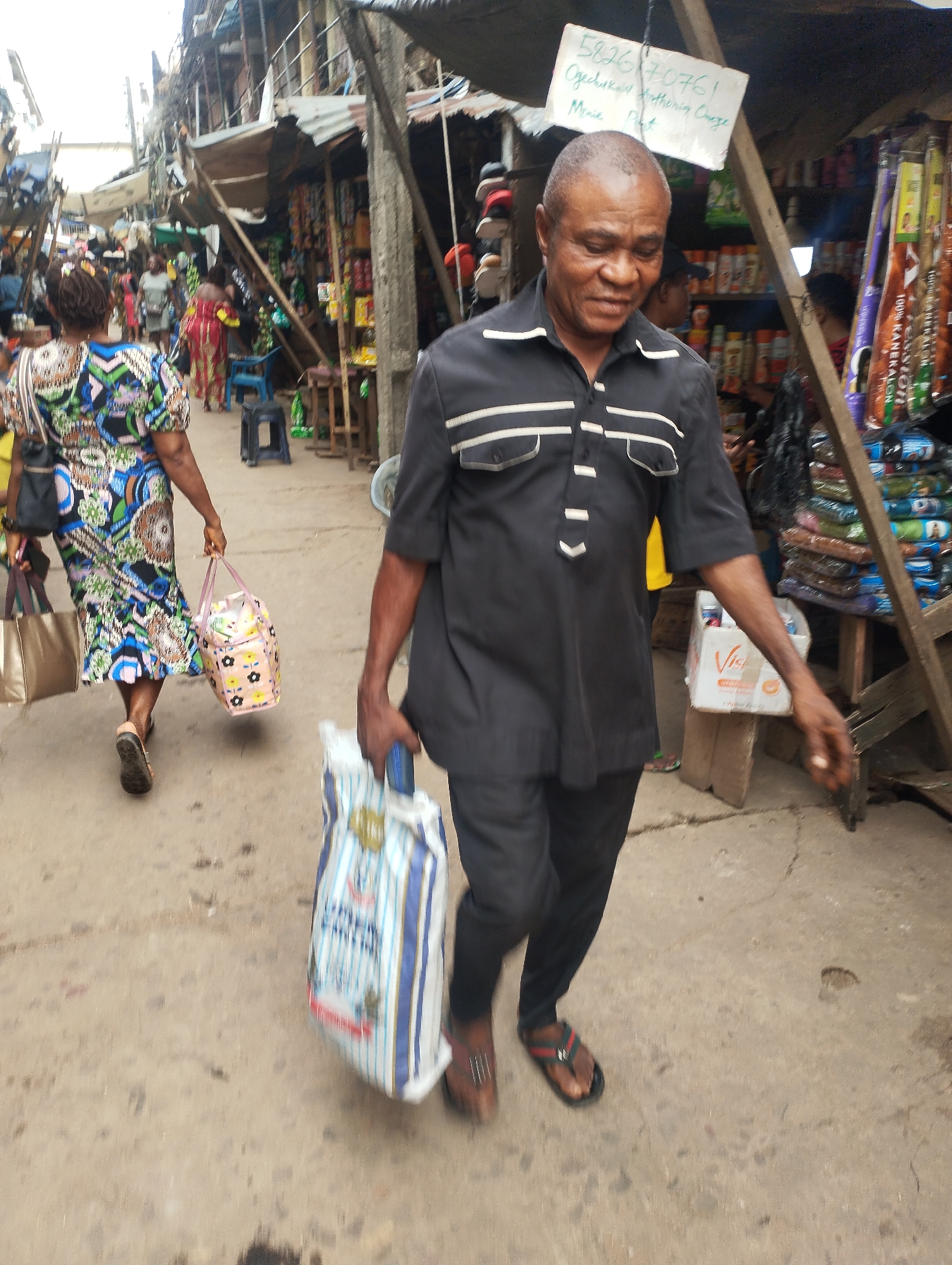 The image depicts a street scene in Fegge, Nigeria. An older man in a black short-sleeved shirt and black pants walks toward the viewer, carrying a large blue-and-white striped bag. He is wearing brown sandals. Behind him, a woman in a colorful patterned dress walks in the same direction, carrying a black purse and a colorful tote bag. The street is lined with stalls selling various goods, and other people are visible in the background. The stalls are covered with makeshift roofs, and the street surface is concrete. A sign with a phone number and name is visible above the stalls.