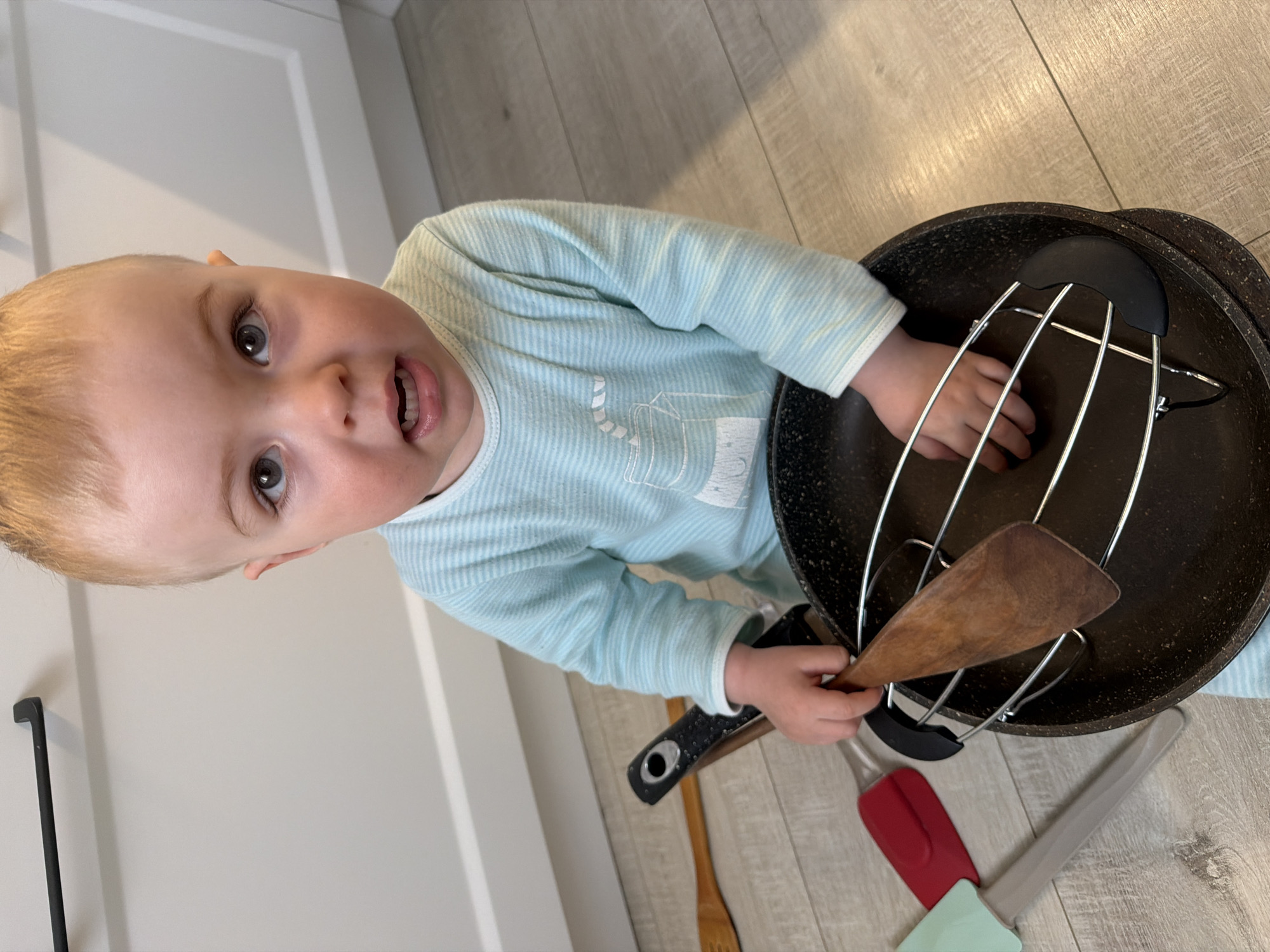A close-up, angled, eye-level shot depicts a young child indoors. The child, wearing a light blue striped long-sleeve shirt with a white graphic on the front, holds a wooden spatula in their right hand. Their left hand is visible near the rim of a black speckled frying pan, which contains a chrome-colored rack. Other kitchen utensils, including a red and gray spatula and a wooden spoon, are on the floor beside the pan. The floor is light-colored woodgrain. A white cabinet is partially visible in the background.
There is no information to indicate that the photograph was taken in Votkinsk, Russia.