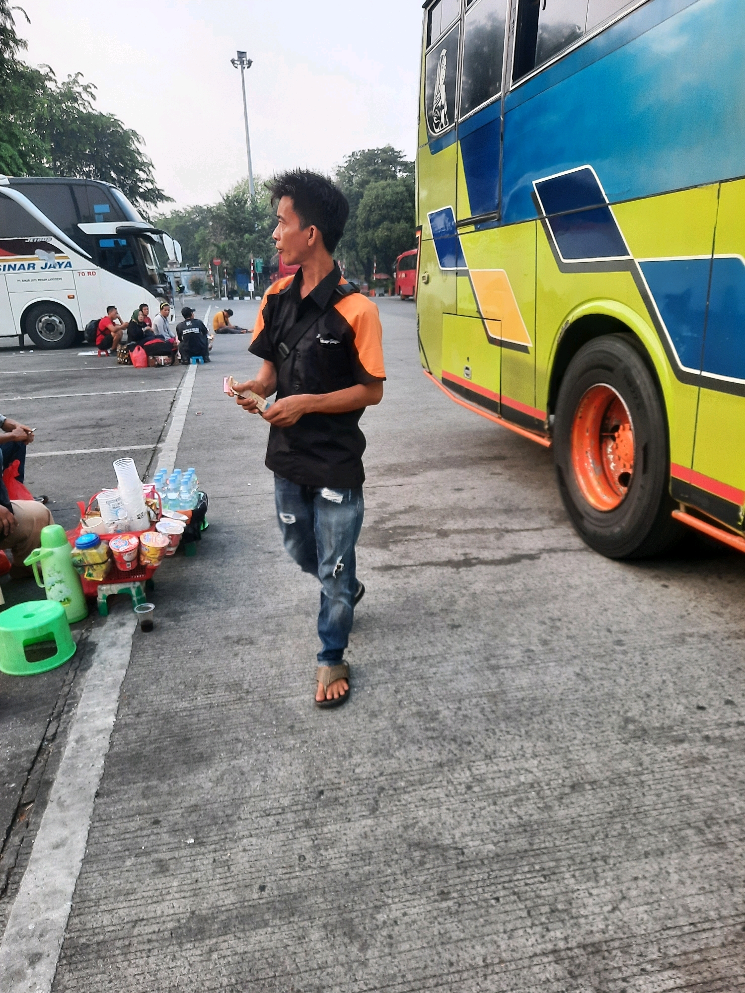 A man in a black shirt with orange sleeves and ripped jeans walks across a concrete surface next to a yellow and blue bus. He is holding some items in his right hand. In the background, a white bus with the text "SINAR JAYA" is parked, and several people are sitting on the ground nearby. A small table with drinks and food items is set up to the left of the man. The scene appears to be a bus station or a rest stop, possibly in East Jakarta, Indonesia, given the context of the buses. The sky is overcast.