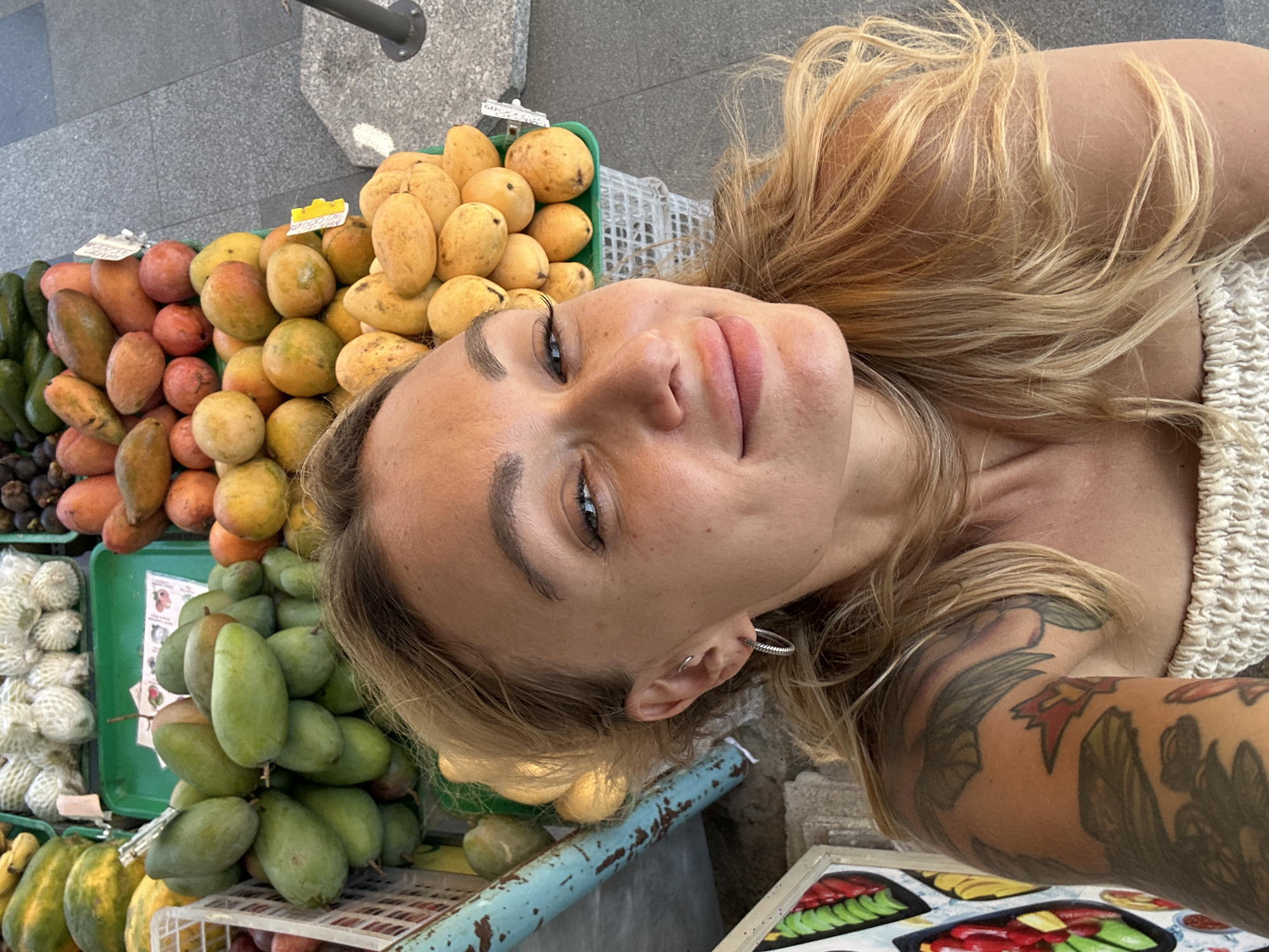 A woman is in Phu Thuy Ward, Vietnam, taking a selfie with a display of fruit at a fruit stand. There are numerous fruits on display, including yellow mangos, green mangos, and other reddish-orange fruits. The woman has blonde hair, tattoos on her arm, and is wearing a light-colored top. The background shows a stone pavement and a glimpse of an object that looks like a metallic post.