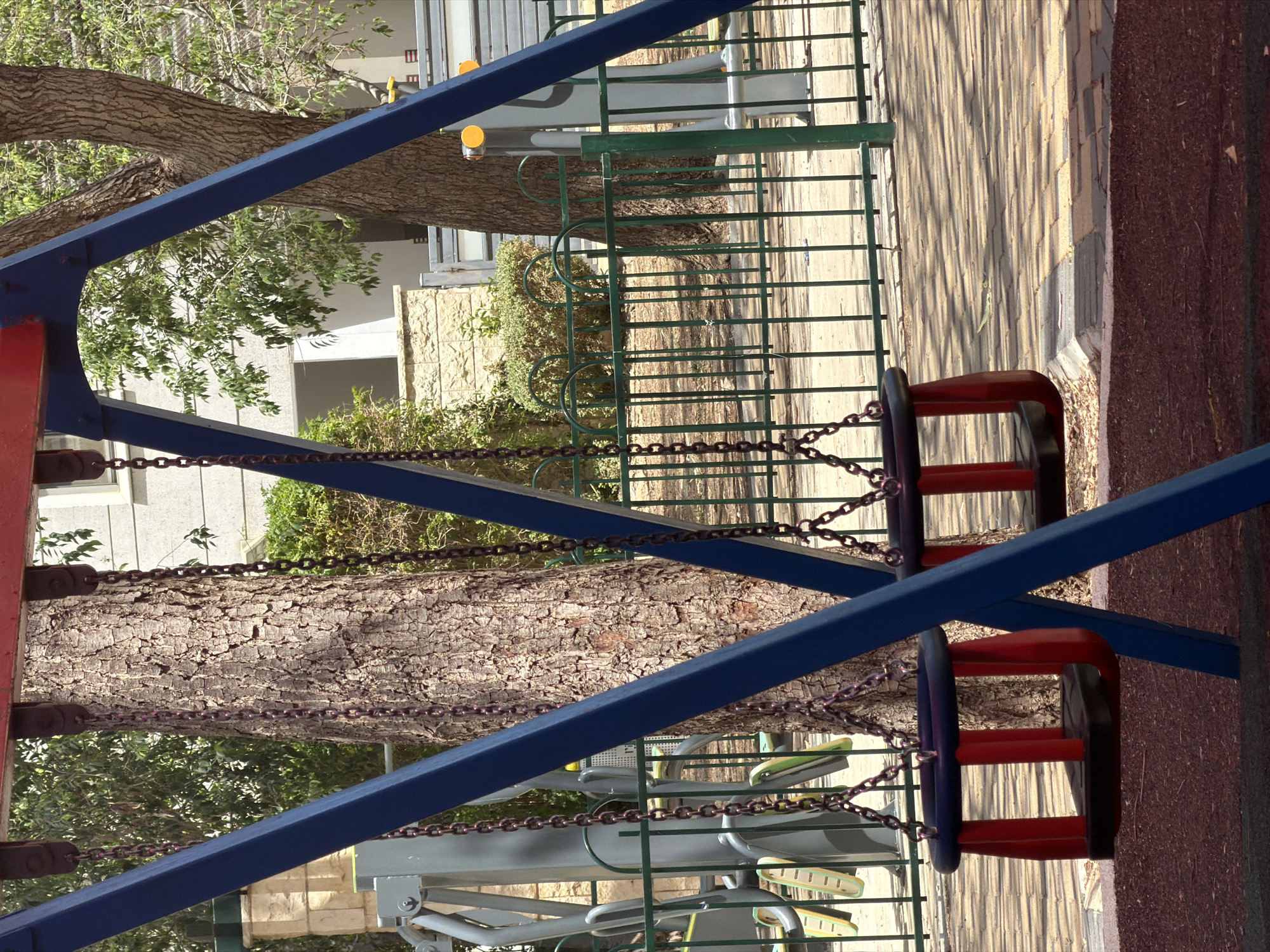 A swing set stands in a playground in Kiryat Motzkin, Israel. The swing set has blue metal support beams with red swing seats hanging from chains. A tree is visible in the background, along with a green fence, outdoor fitness equipment, and a building. The ground appears to be paved and covered with rubberized material. The image is rotated 90 degrees counterclockwise.