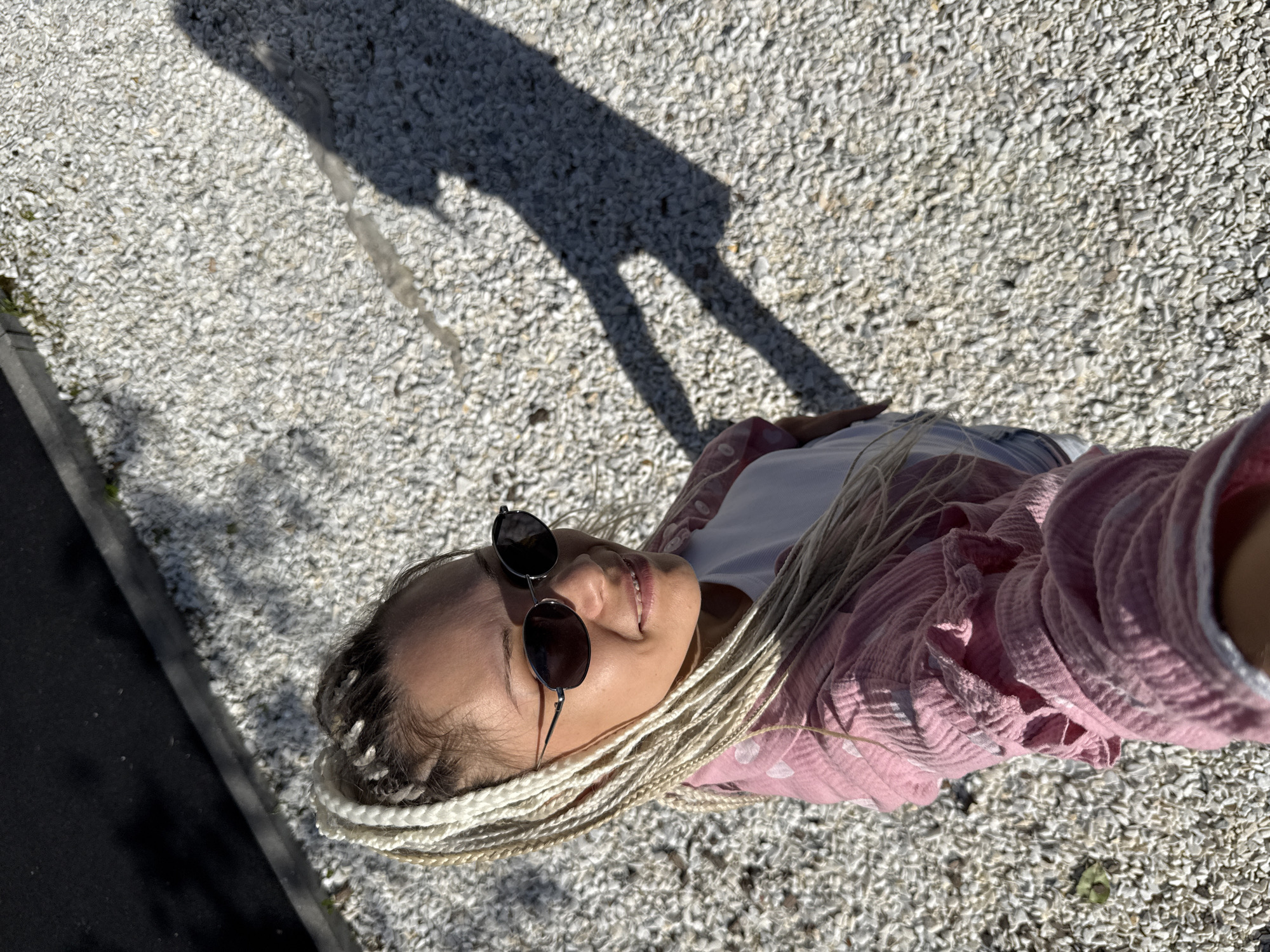 A high-angle selfie shows a woman with blonde braids wearing sunglasses and a pink button-up shirt over a white t-shirt. She is standing on a gravel path, with a dark, shadowed area on the left side of the frame that appears to be a wall or sidewalk. The sun casts a long shadow of the woman to her right, which is also visible on the gravel. The overall setting is outdoors, likely in a public space or park, with a light gravel ground cover. There is no indication of the specific location within Rusanovo, Russia.