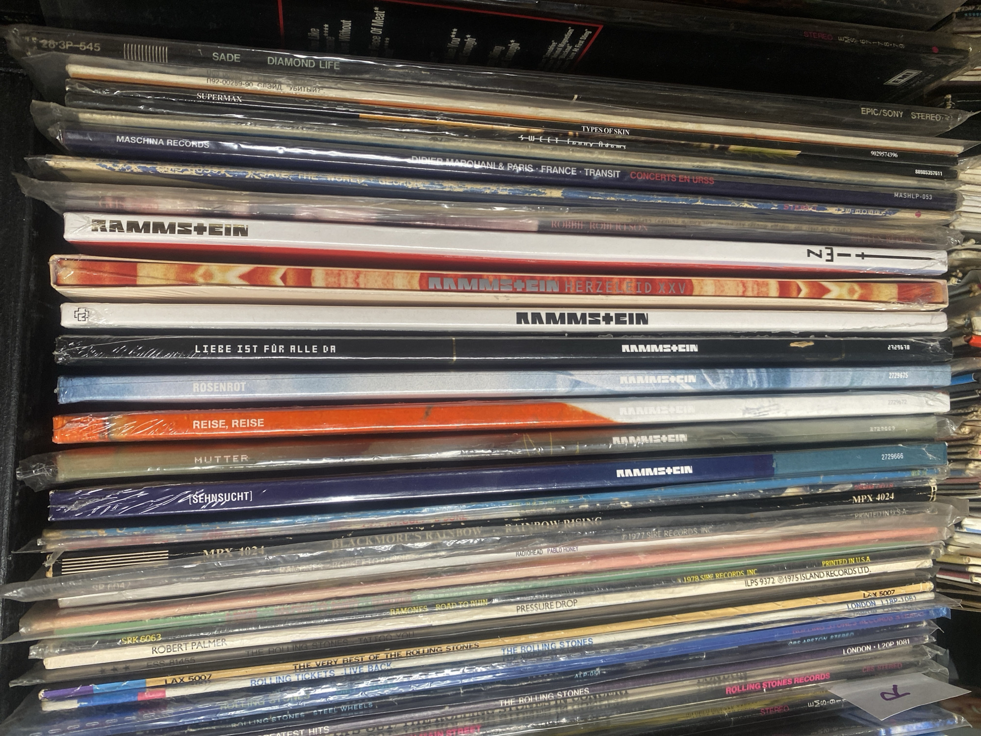 The image shows a close-up, vertical view of a shelf filled with vinyl records. The records are tightly packed, with many of them still in their plastic sleeves, creating a slightly distorted and reflective surface. The spines of the records are the primary focus, displaying various album titles and artist names in different fonts and colors. Some of the visible text includes "RAMMSTEIN," "REISE, REISE," "MUTTER," "ROSENROT," "LIEBE IST FÜR ALLE DA," "DIAMOND LIFE," and "ROLLING STONES." The overall impression is of a music collection, likely stored in a home or a record store. There are no people visible in the image. The environment appears to be indoors, with artificial lighting. The specific location of Lindo-Pustyn' (Kantaurovskii S/S), Russia, is not depicted or suggested by any visual cues in the image.